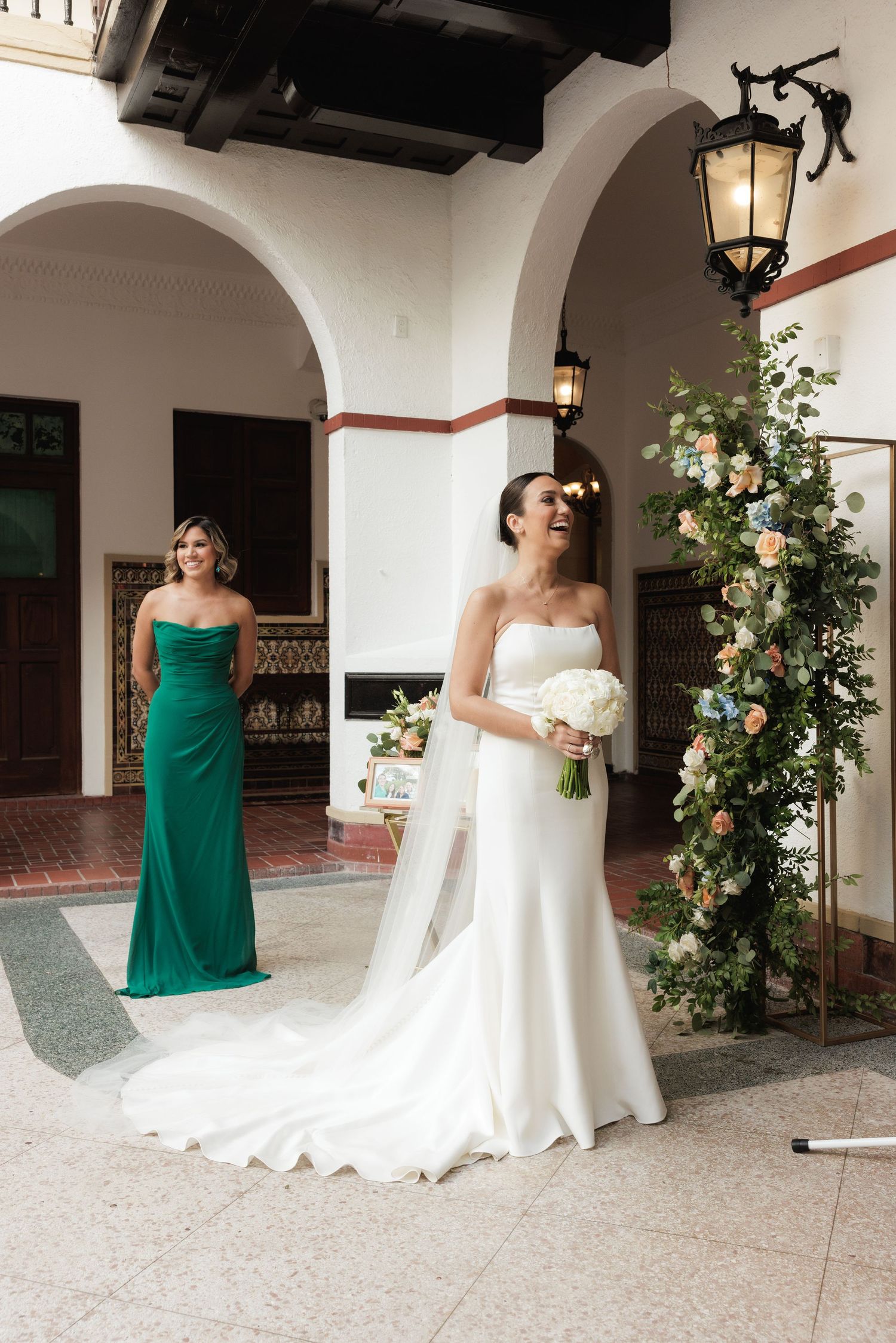 Wedding party poses in elegant Spanish-style courtyard with white arches and floral arrangements.