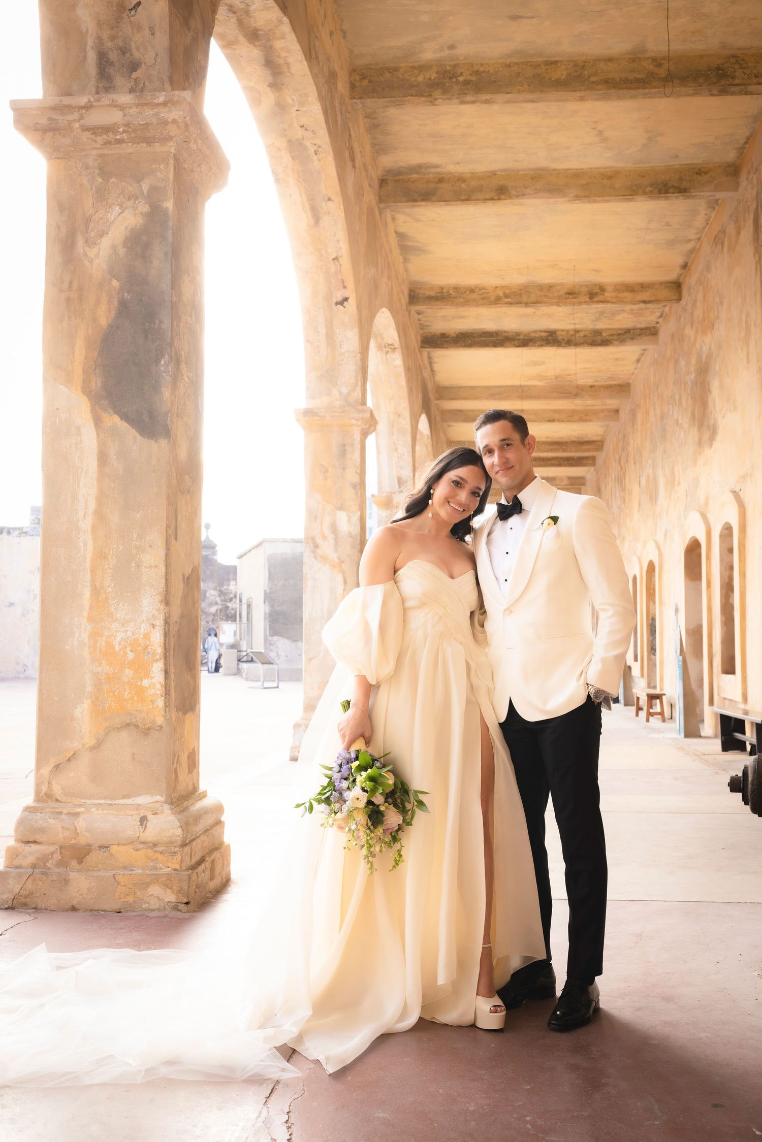 Wedding couple poses under historic stone archway colonnade in romantic outdoor setting.