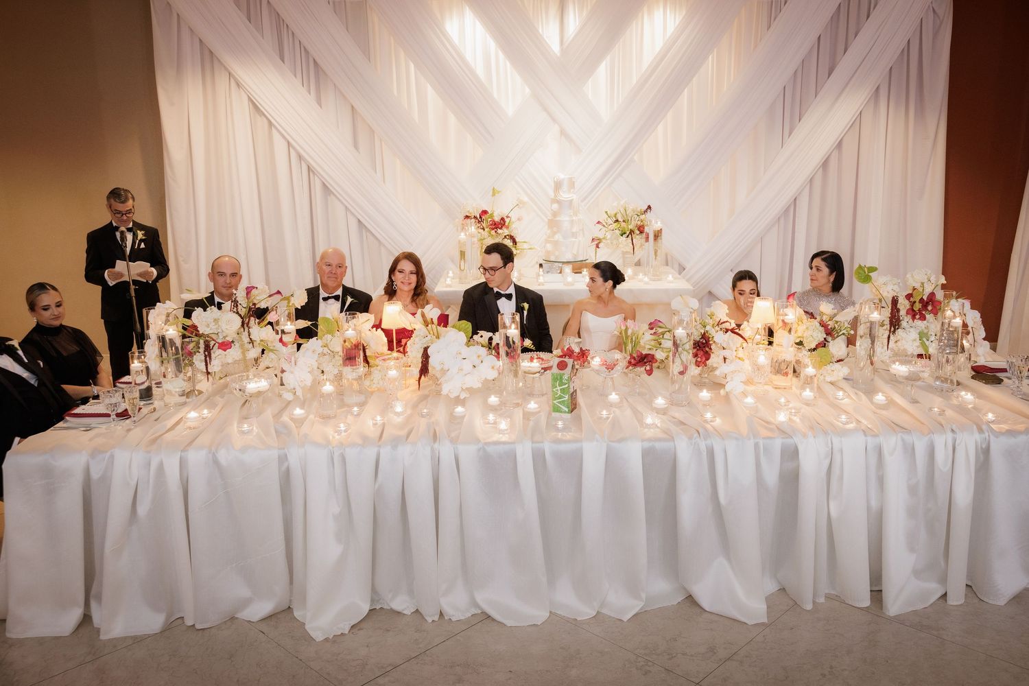 A long white wedding reception head table decorated with elegant floral centerpieces and glowing candlelight.