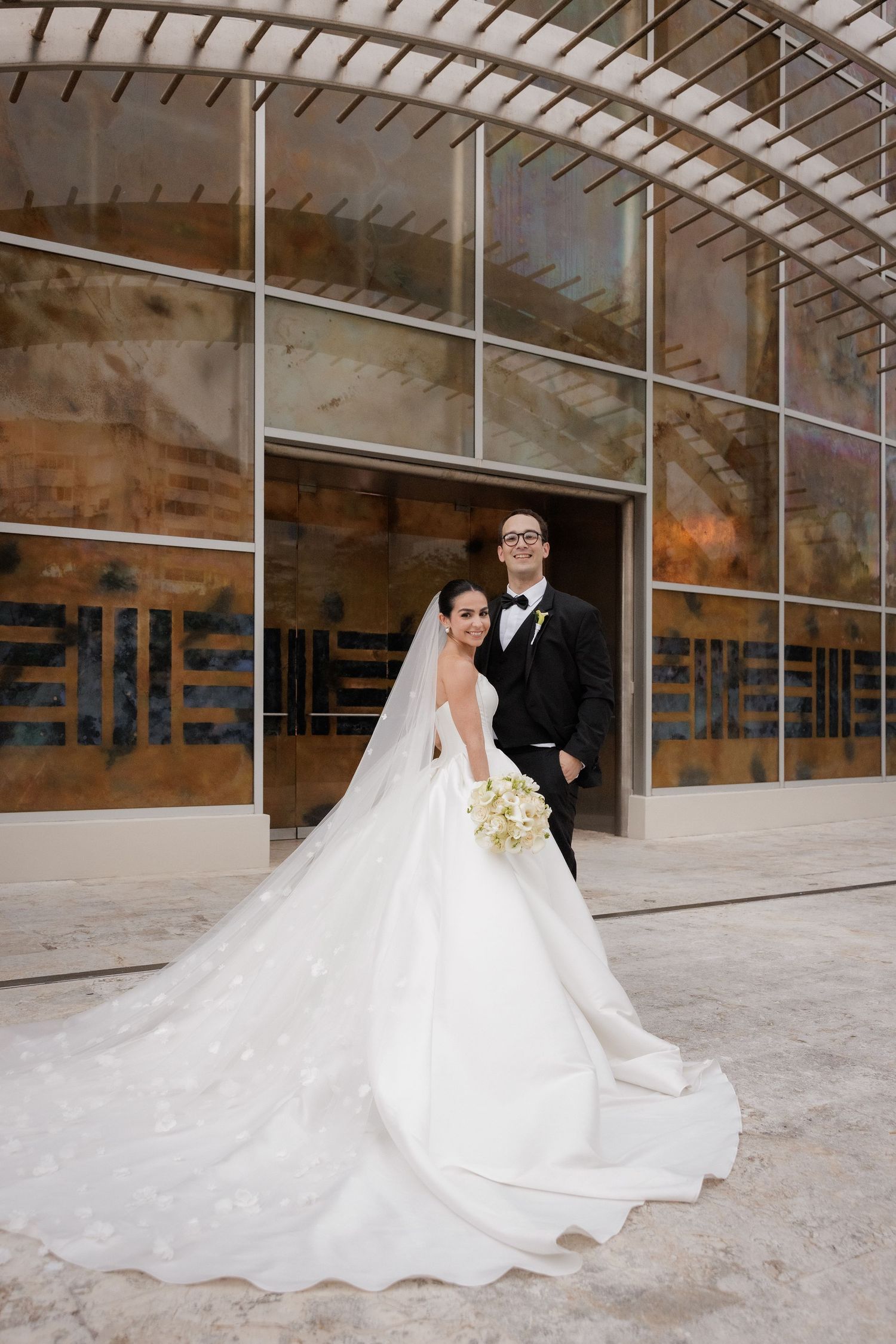 A bride and groom pose in front of a modern architectural building with glass windows and a flowing white wedding dress.