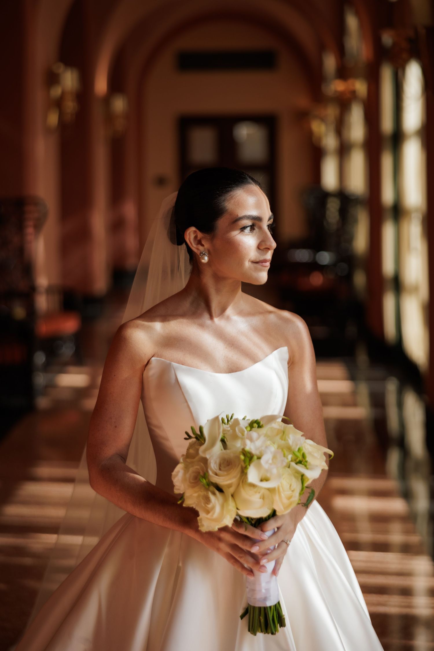 A bride in a strapless white gown holds a bouquet of white flowers while standing in an elegant hallway with arched windows.