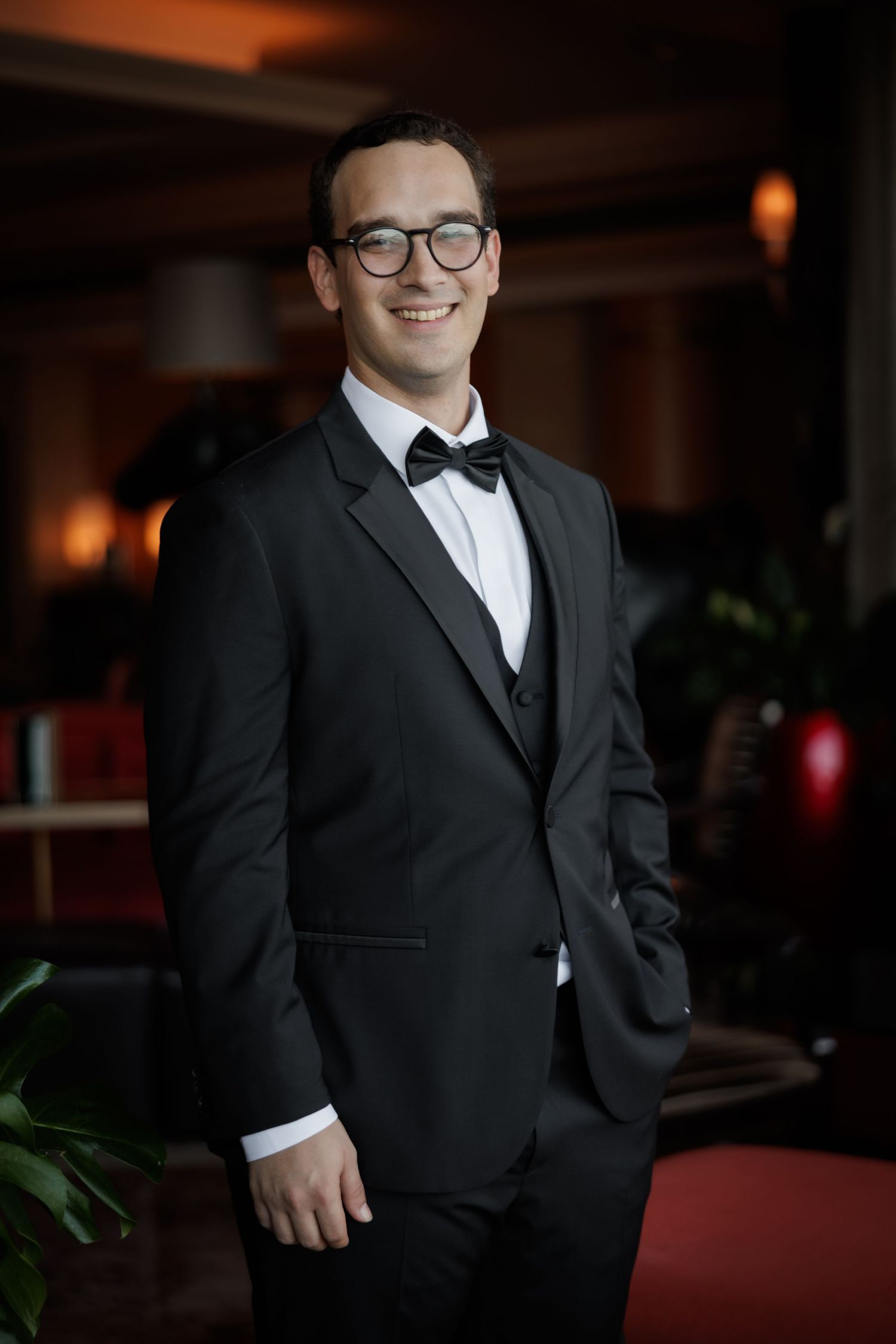 A groom in a classic black tuxedo and bow tie smiles in a dimly lit room.