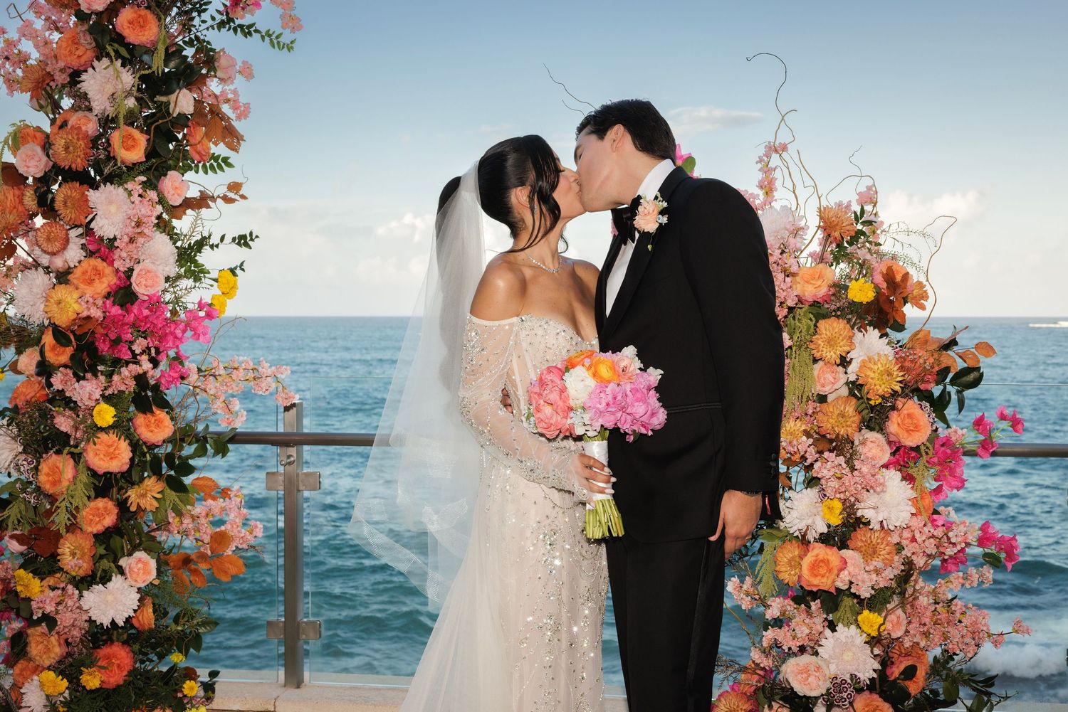 Romantic wedding ceremony by the ocean with a floral arch of pink and peach roses framing a newlywed couple kissing.