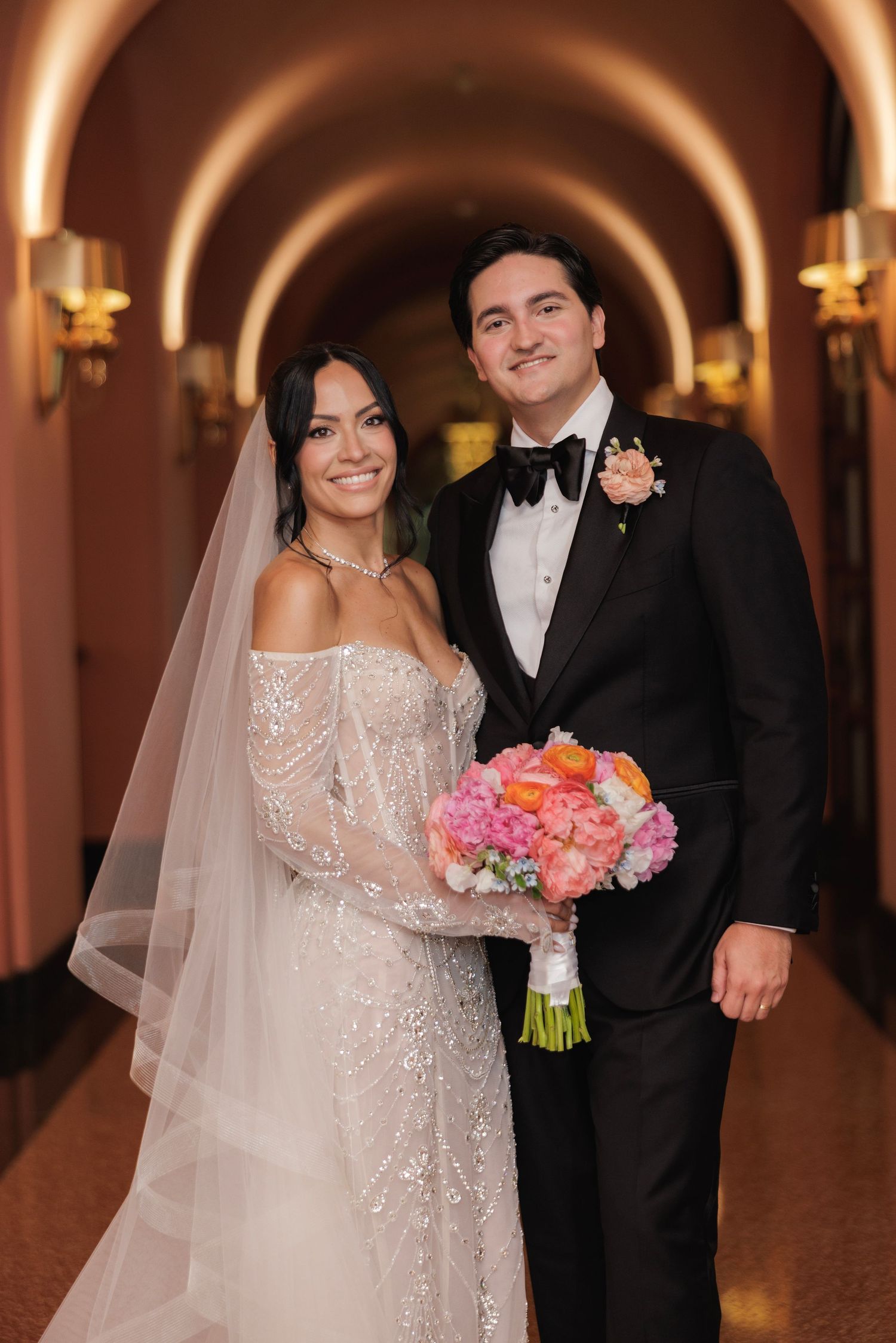 A bride in an off-shoulder lace gown and groom in a black tuxedo pose together in an elegant arched hallway.
