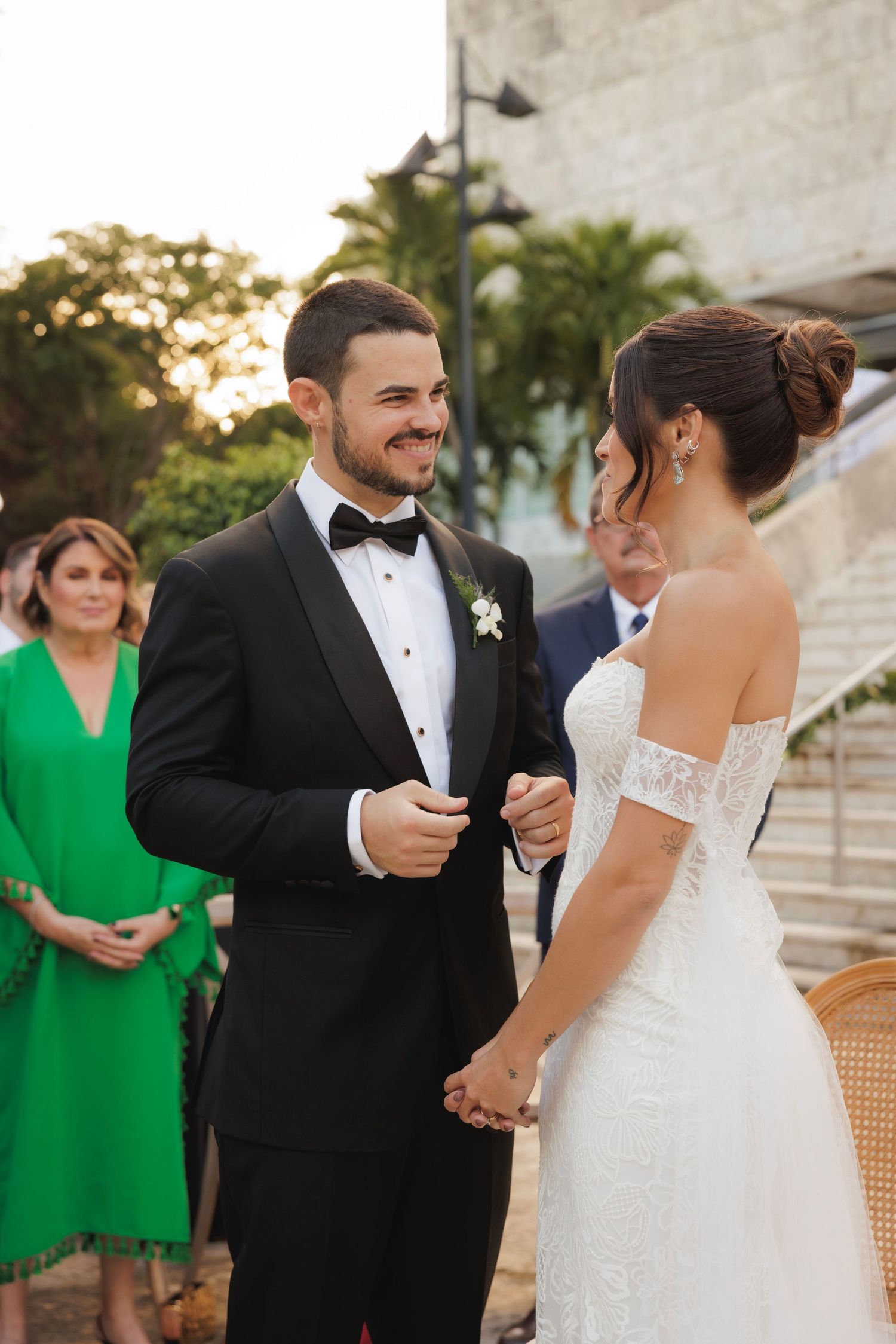 Elegant outdoor wedding scene with white lace dress and black tuxedo against stone architecture.
