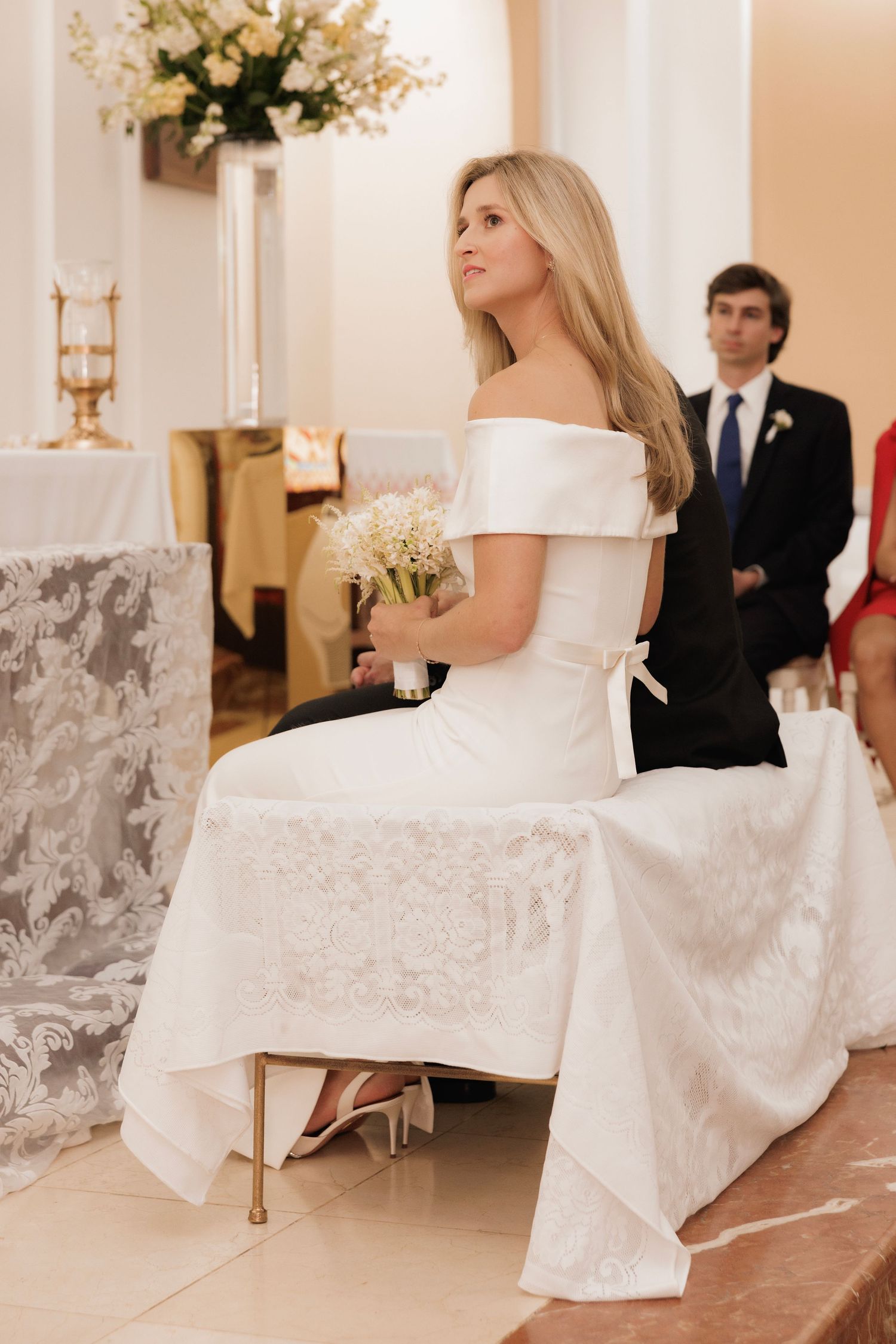 A bride in a white wedding dress sits elegantly on a gold chair in front of a soft neutral wall during a wedding event.