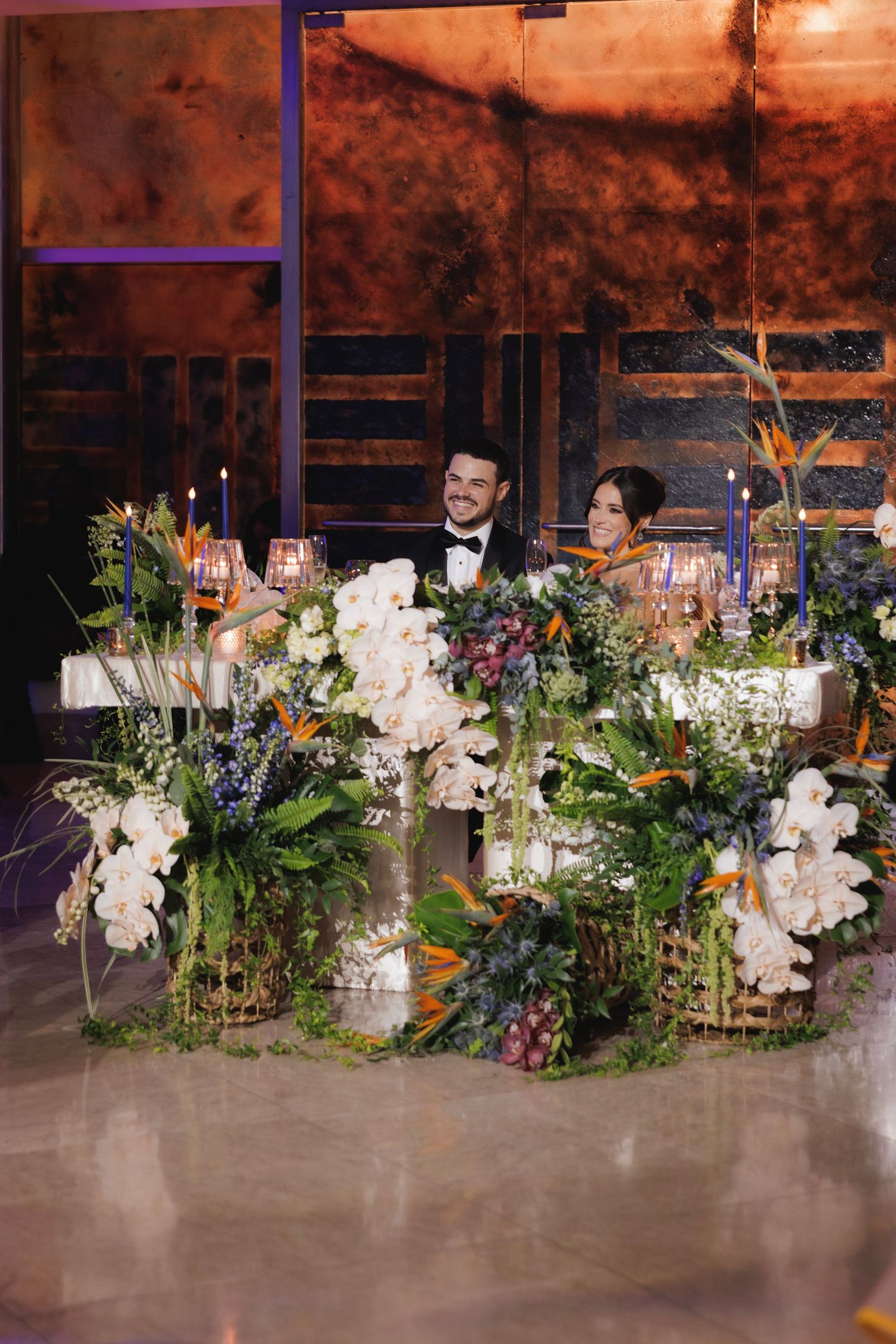 Elegant floral table arrangements featuring white orchids and mixed greenery line a wedding reception space.