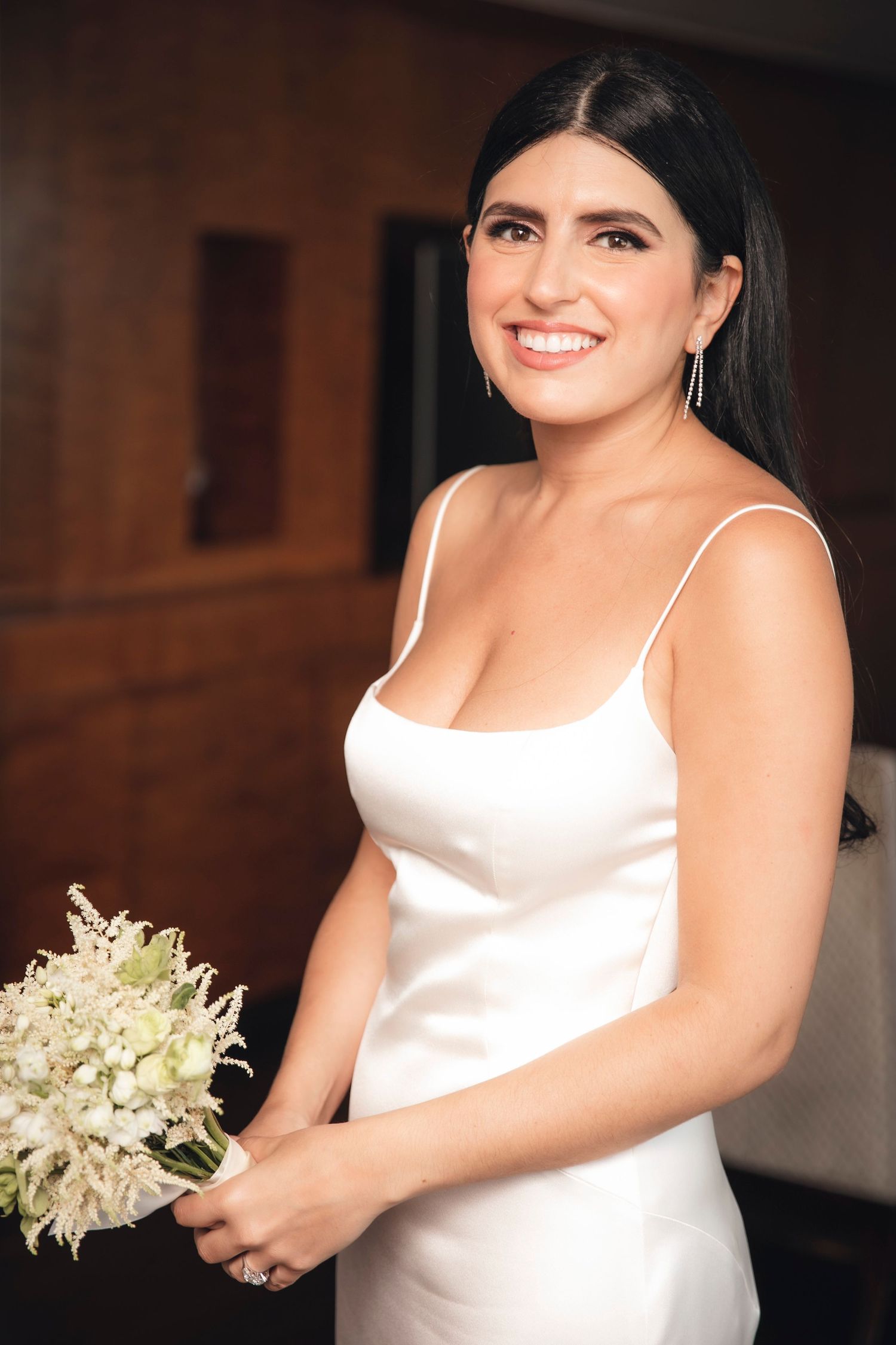 Smiling woman in simple white satin wedding dress holds delicate bouquet of white flowers against dark interior.