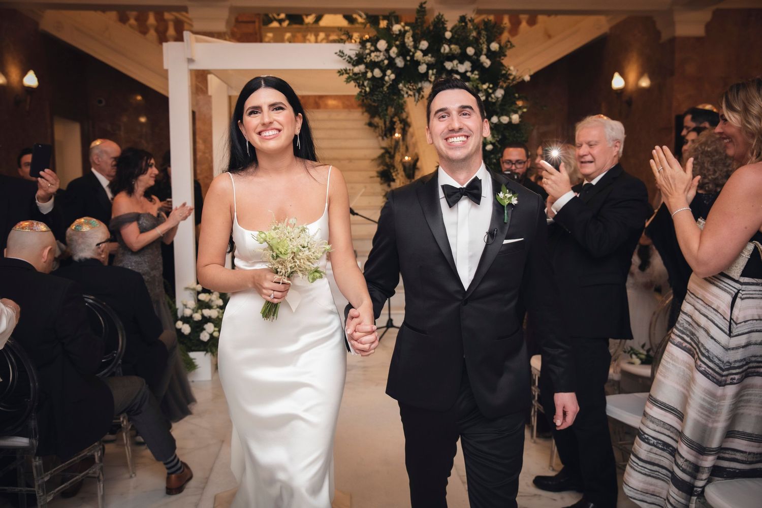 Guests dance and celebrate at an elegant indoor wedding reception with warm lighting and decorative greenery.