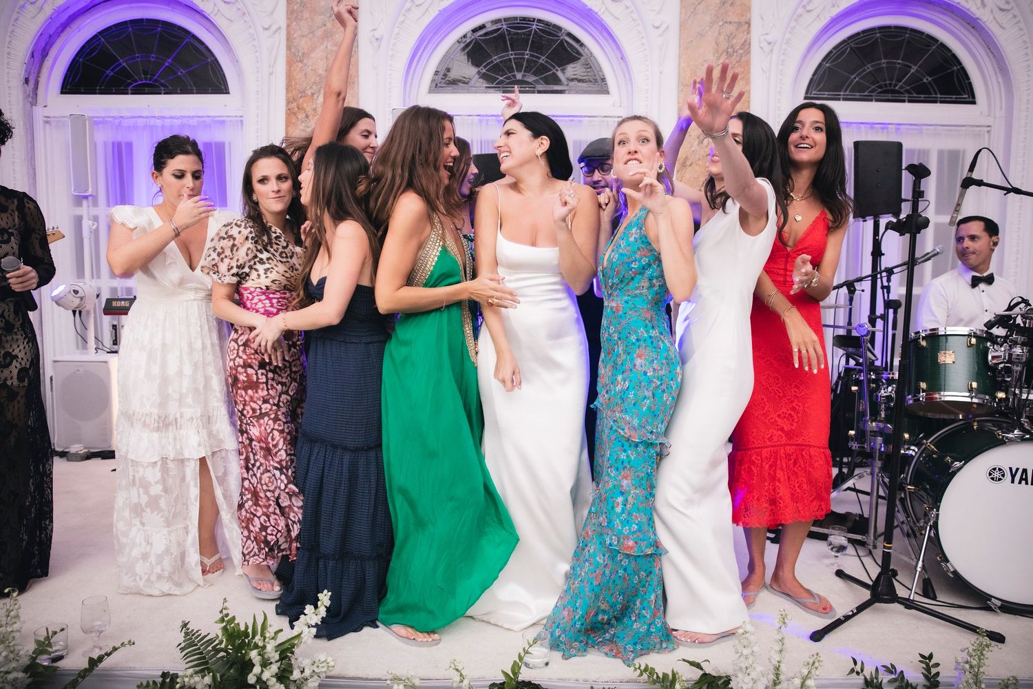 Wedding party and guests dancing together in an elegant ballroom with purple-lit archways.
