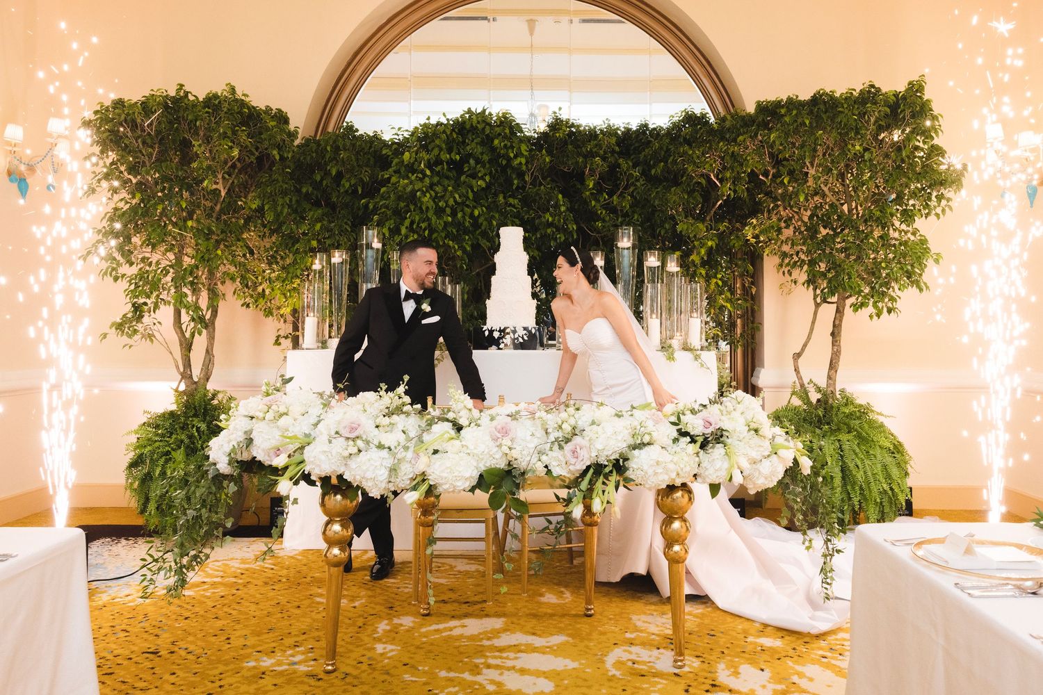 Elegant wedding reception table setting with white floral centerpieces and tall greenery under an ornate archway.