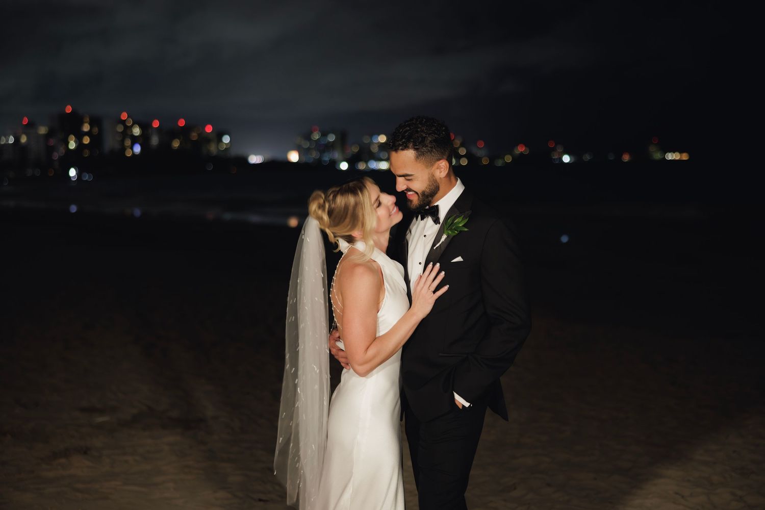 A couple shares a romantic kiss at night during their wedding celebration with soft lighting in the background.