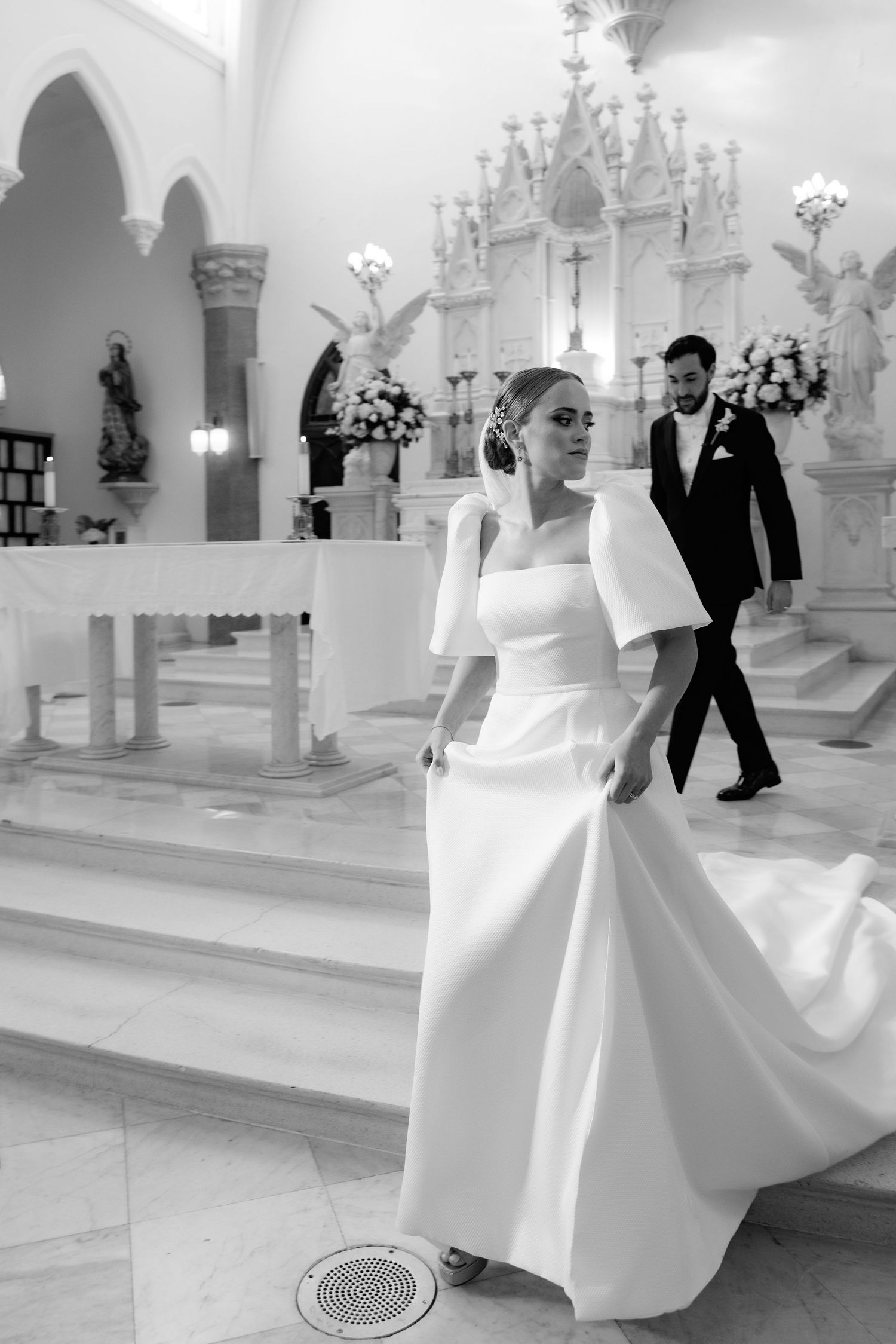 Elegant wedding ceremony in a church interior with ornate white altar and architectural arches in black and white.