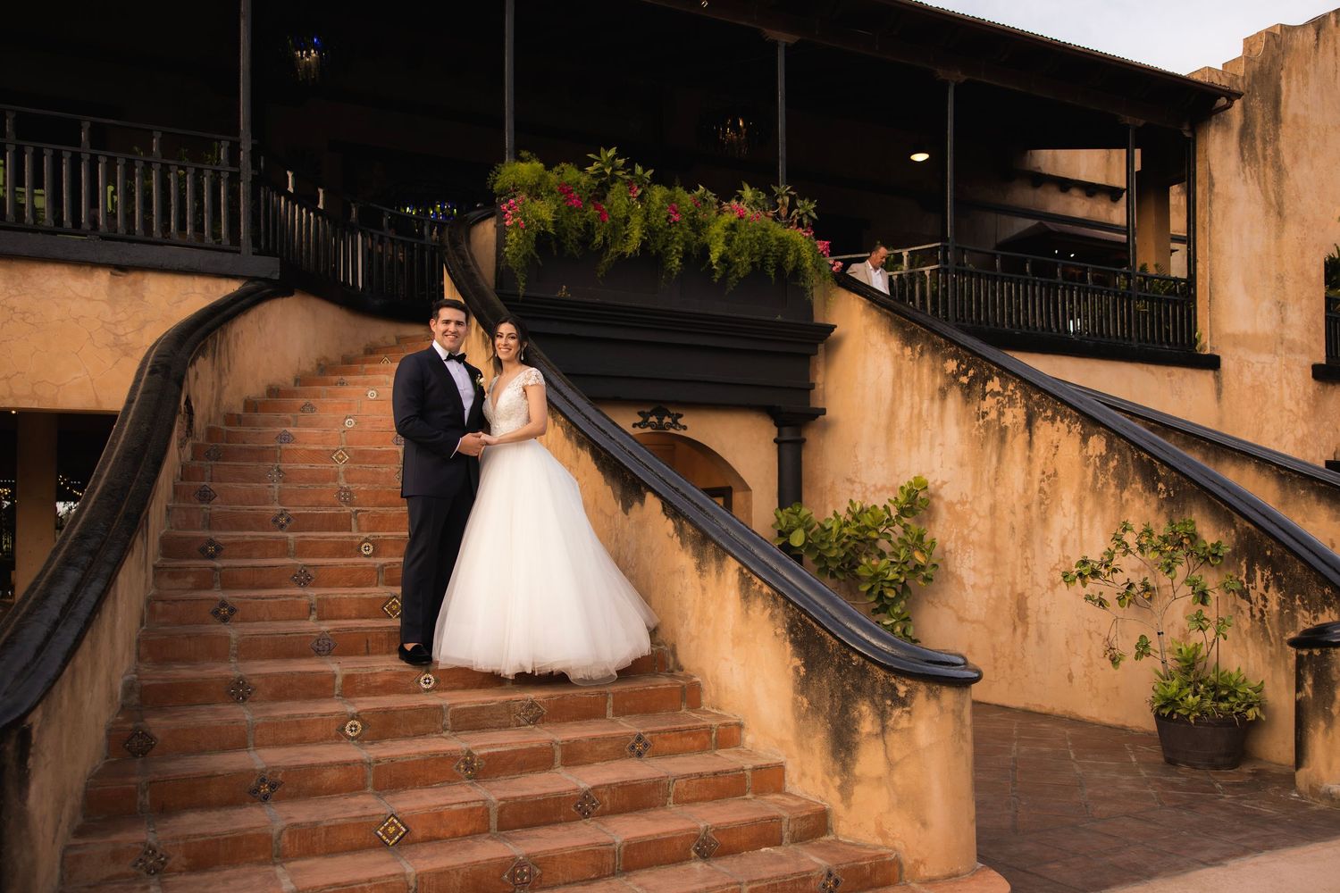 Bride in flowing white gown and groom ascend rustic stone staircase during sunset photo session.