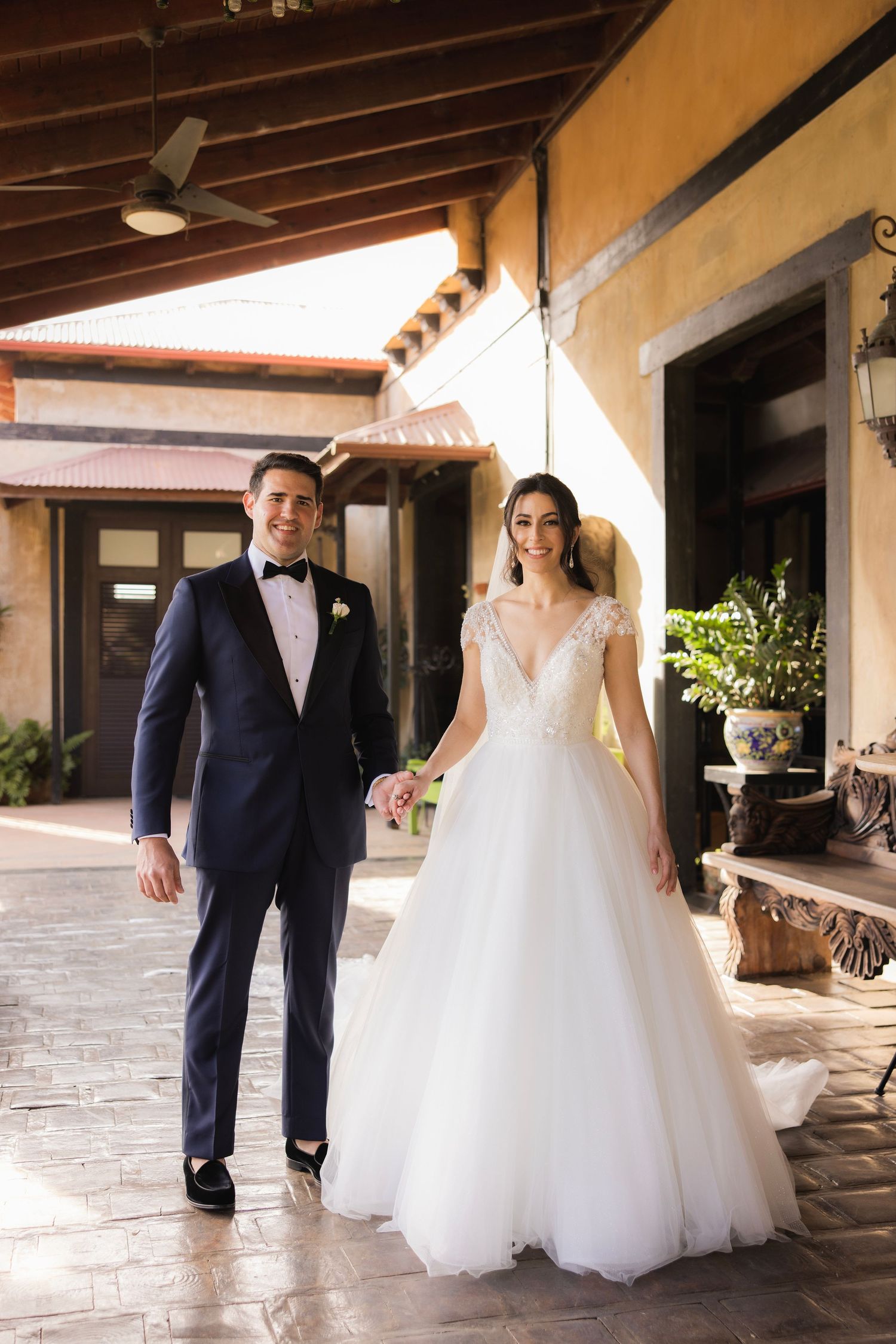 A couple shares a joyful moment walking together through an elegant covered walkway in their wedding attire.