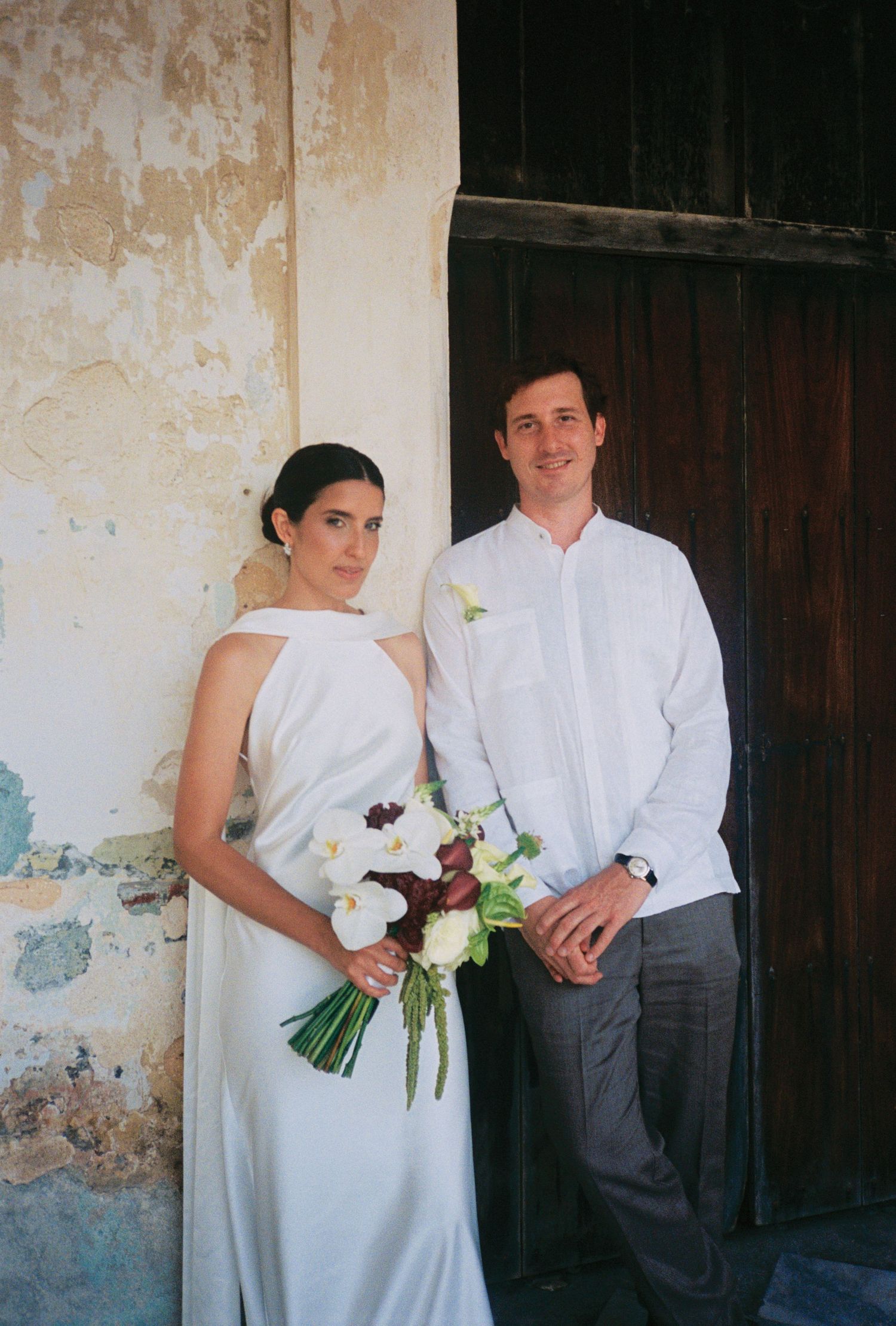 Bride in white halter gown holding floral bouquet stands beside groom in white guayabera shirt near rustic wall.
