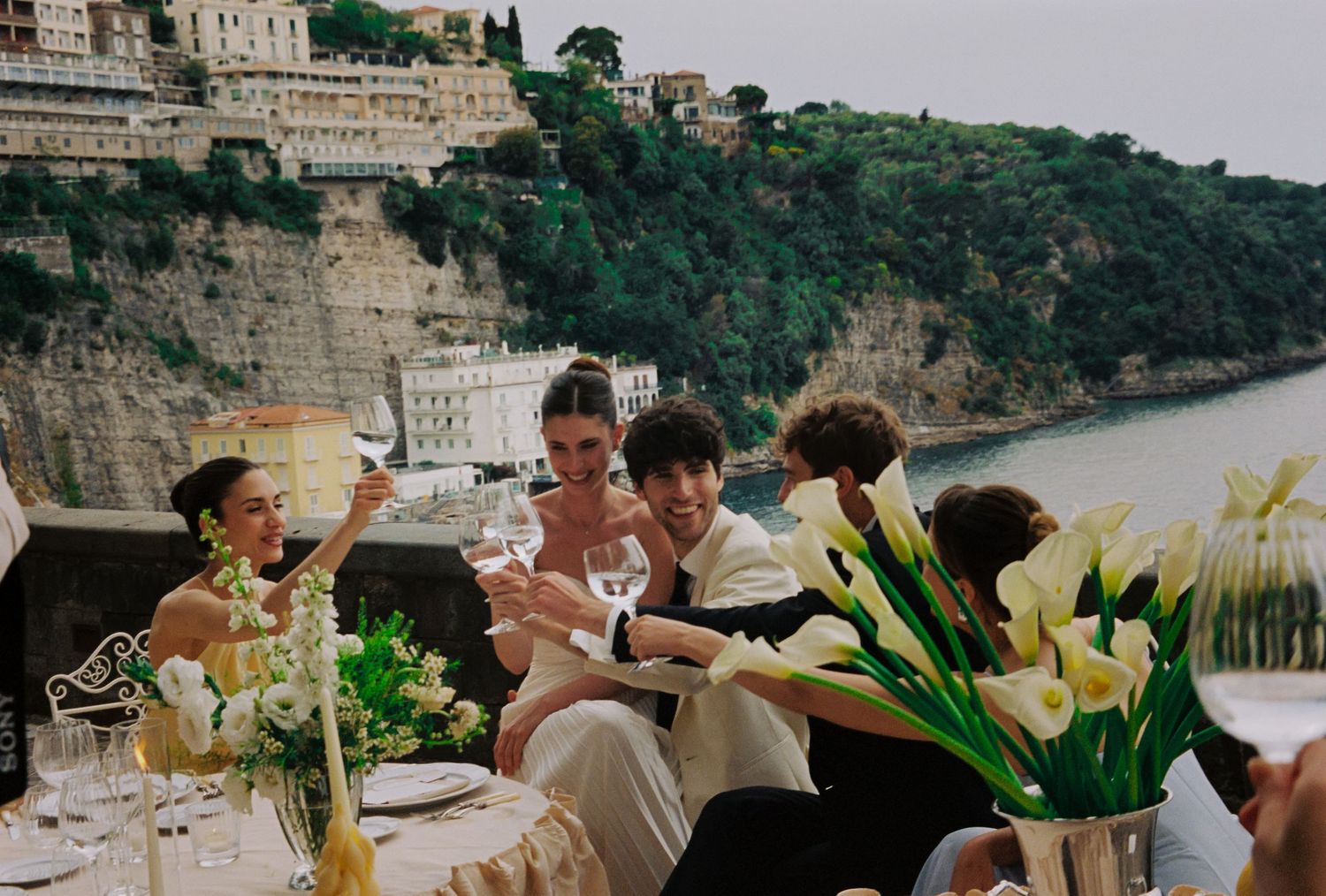 Elegant outdoor wedding reception with guests toasting on a cliffside terrace overlooking the sea in Amalfi Coast, Italy.