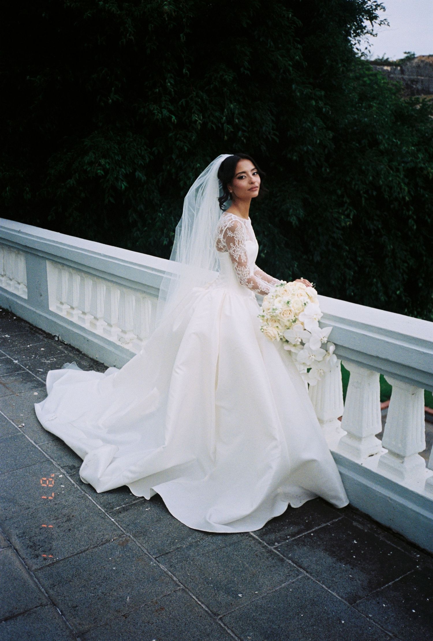 Elegant bride in a white ball gown with lace sleeves and veil holding a bouquet on a balcony.
