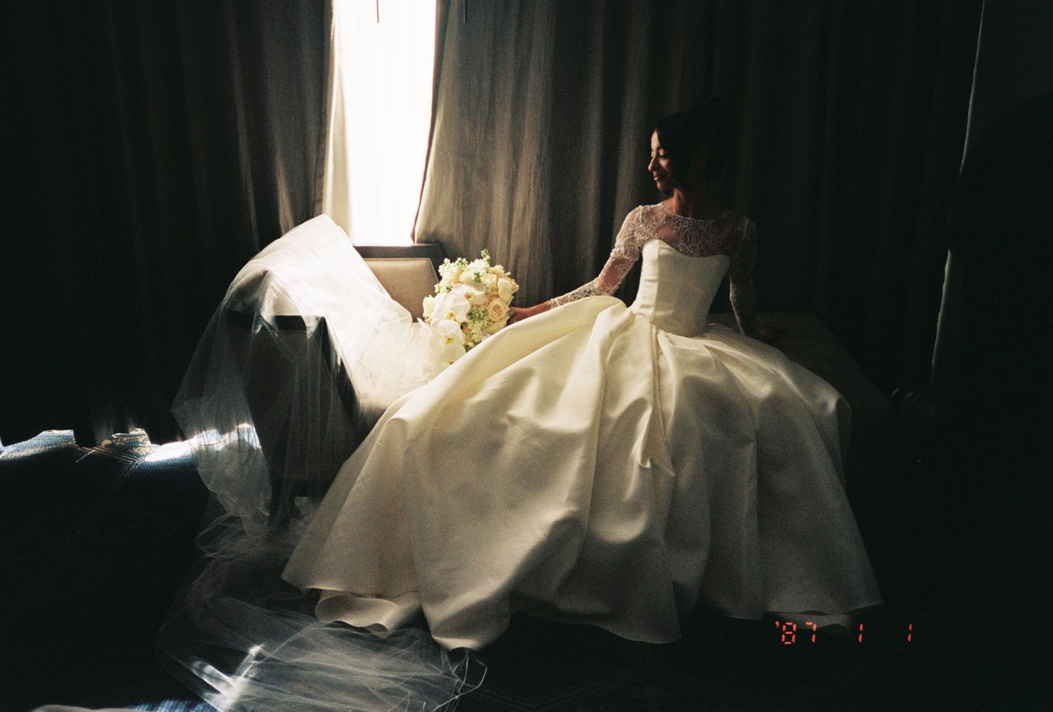 Bride in elegant white ball gown wedding dress holding bouquet, seated by window in soft natural light.