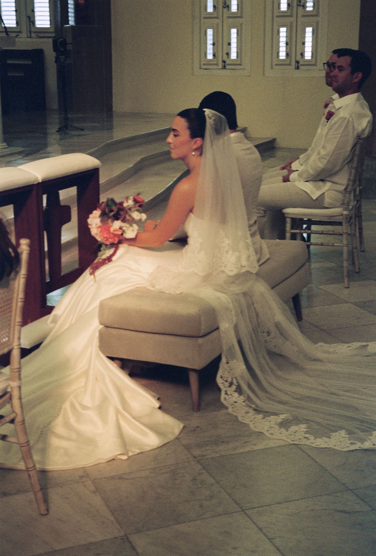 Bride in lace-trimmed veil and white gown seated during church ceremony with groom beside her.