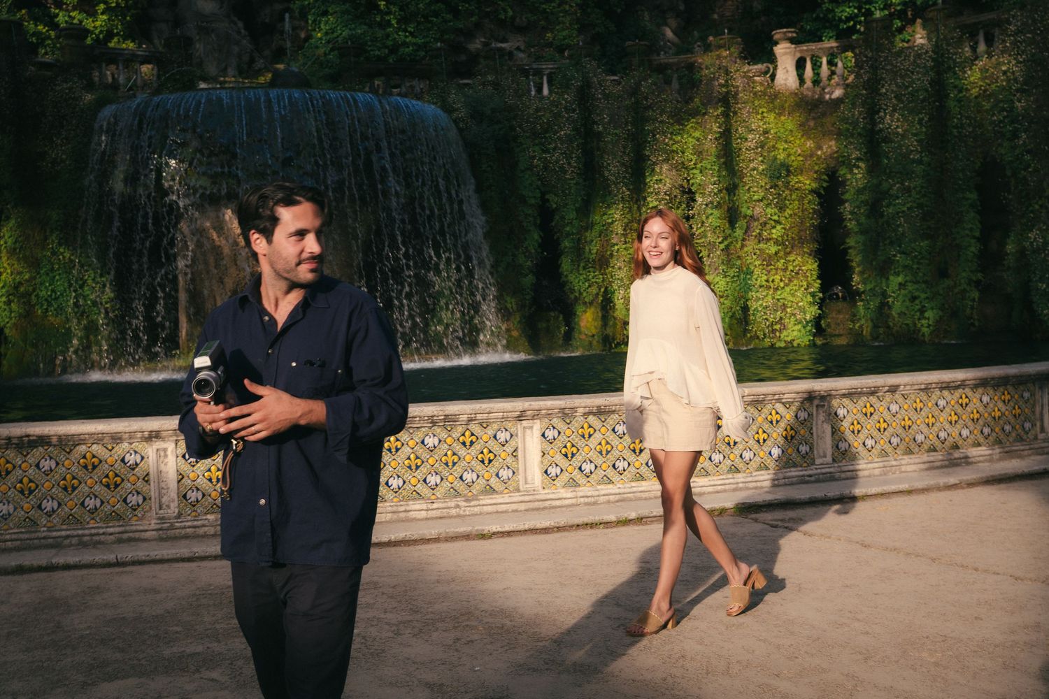 A man holding a camera and a woman in a white dress pose near an ornate fountain surrounded by lush greenery.