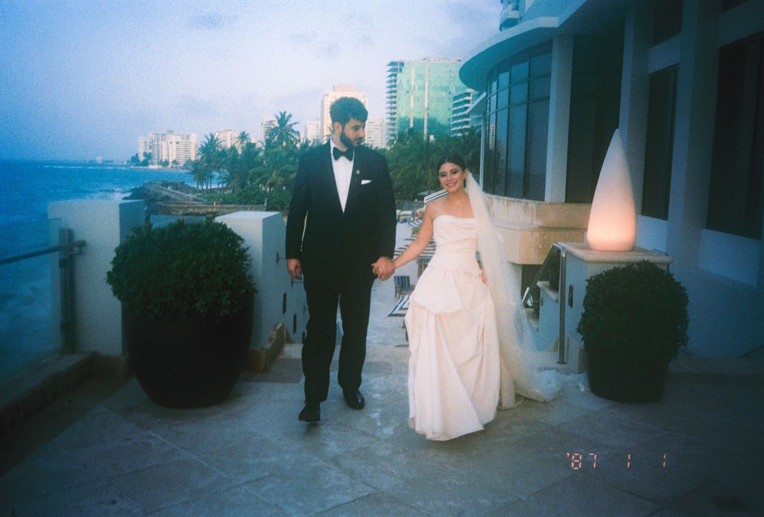 Bride and groom hold hands on oceanfront terrace at dusk, she in white gown, he in black tuxedo.
