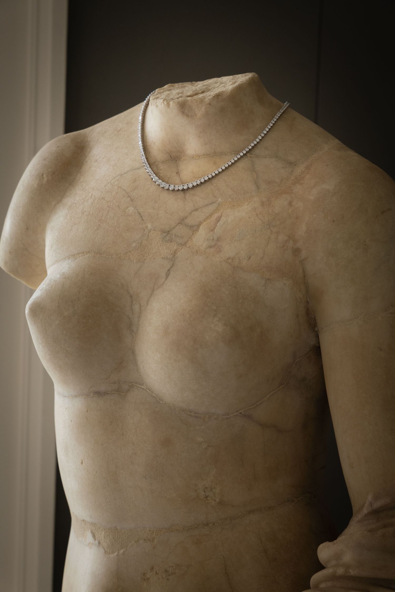 Ancient marble torso sculpture adorned with a delicate silver diamond tennis necklace displayed against a dark background.