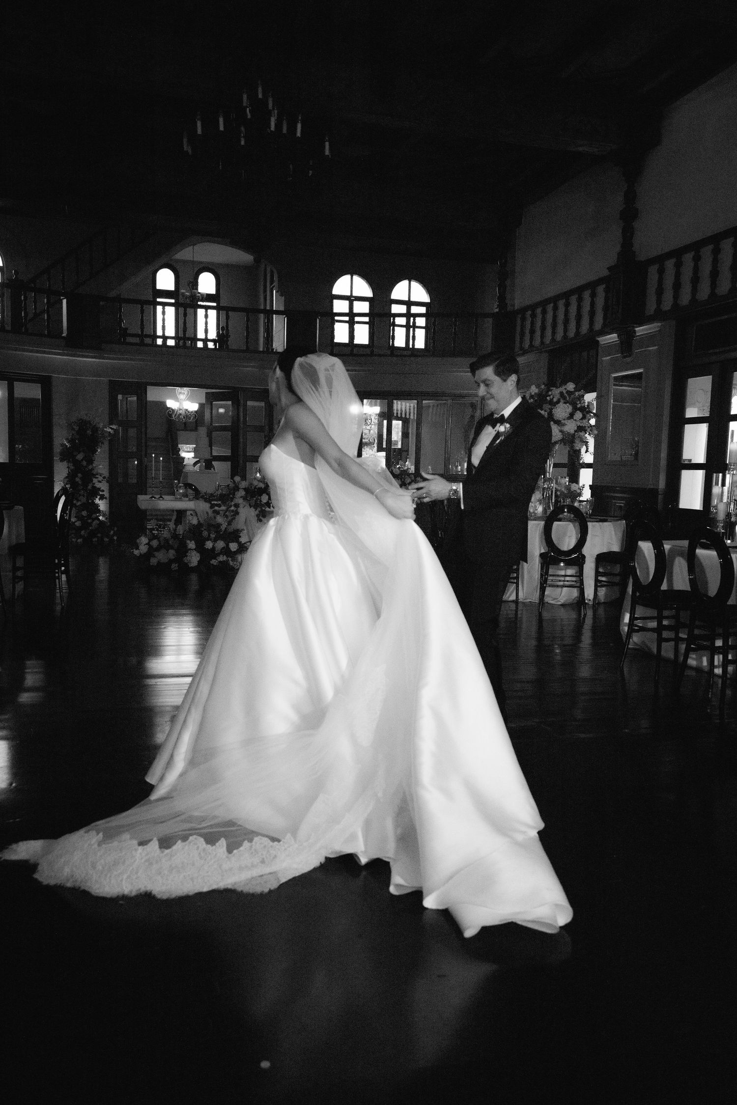Silhouetted wedding couple dancing in a dimly lit ballroom with arched windows creating a dramatic atmosphere.