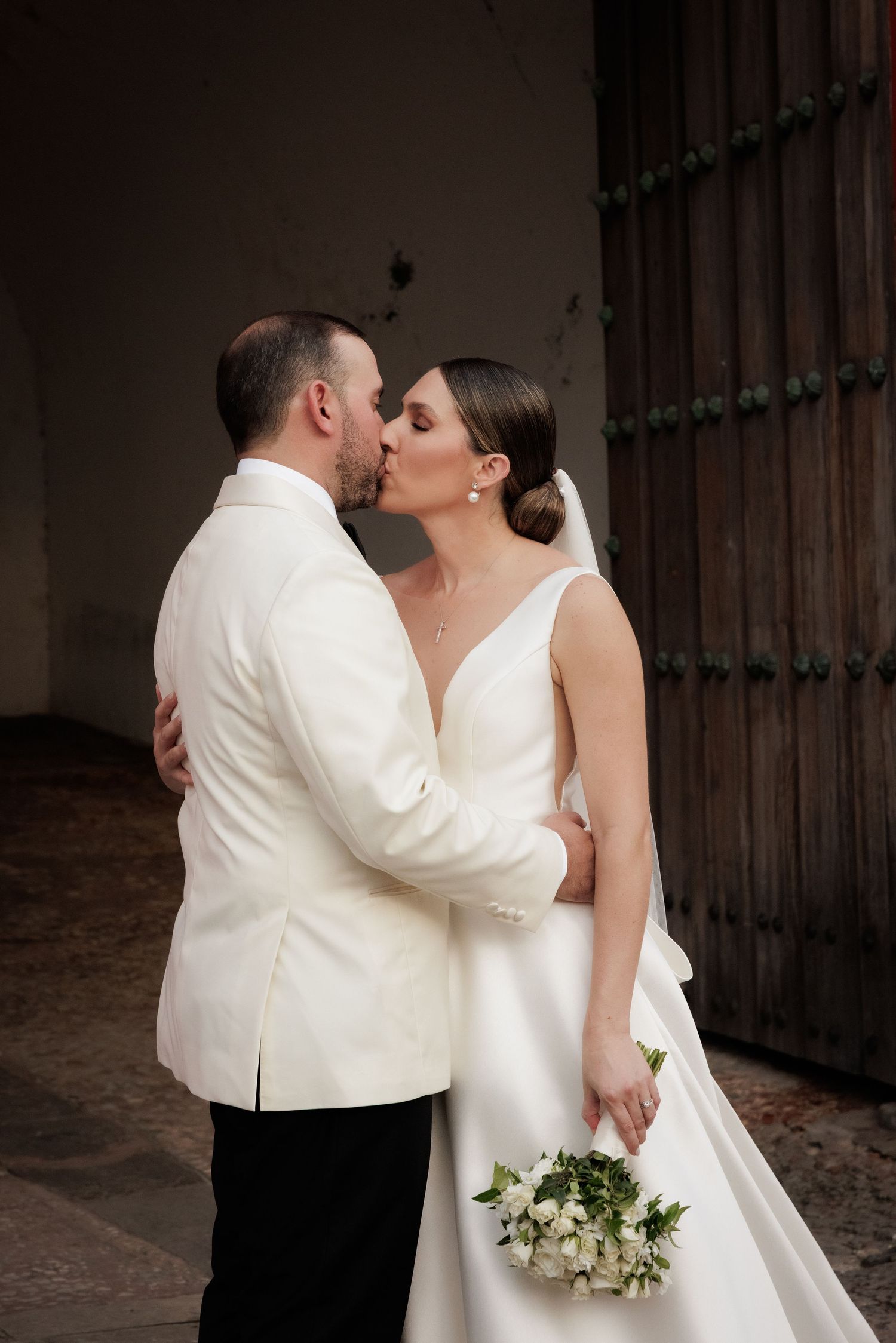 A romantic wedding moment captured as the couple shares a kiss against an ornate wooden door backdrop.