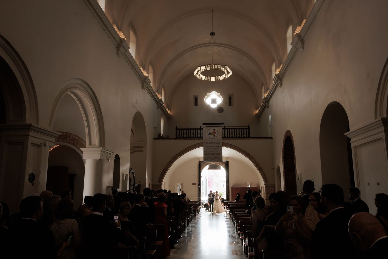 Silhouetted figures stand in a dimly lit church interior with arched ceilings and dramatic backlighting through doorways.