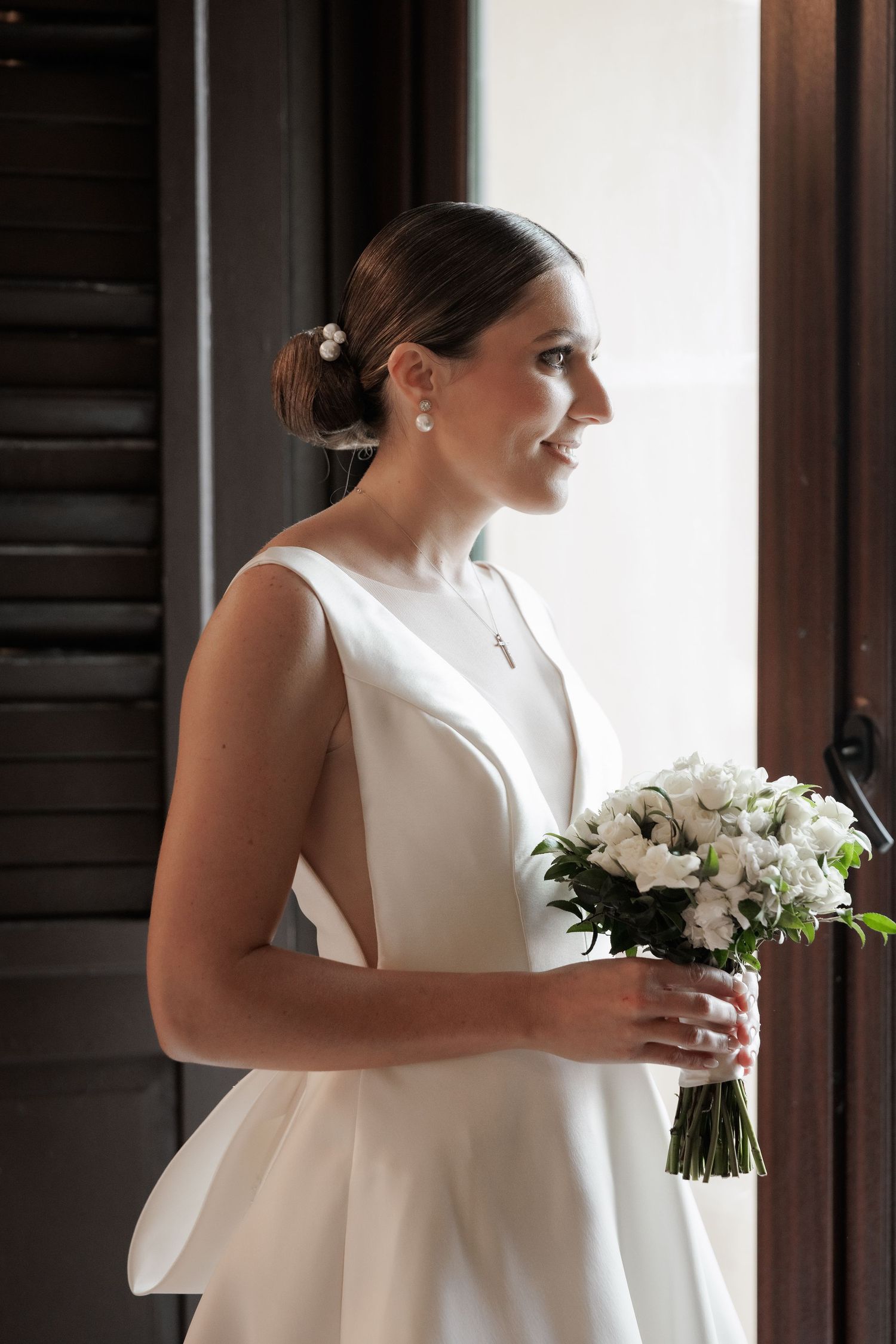 A person in an elegant white wedding dress holds a bouquet of white flowers near a window.