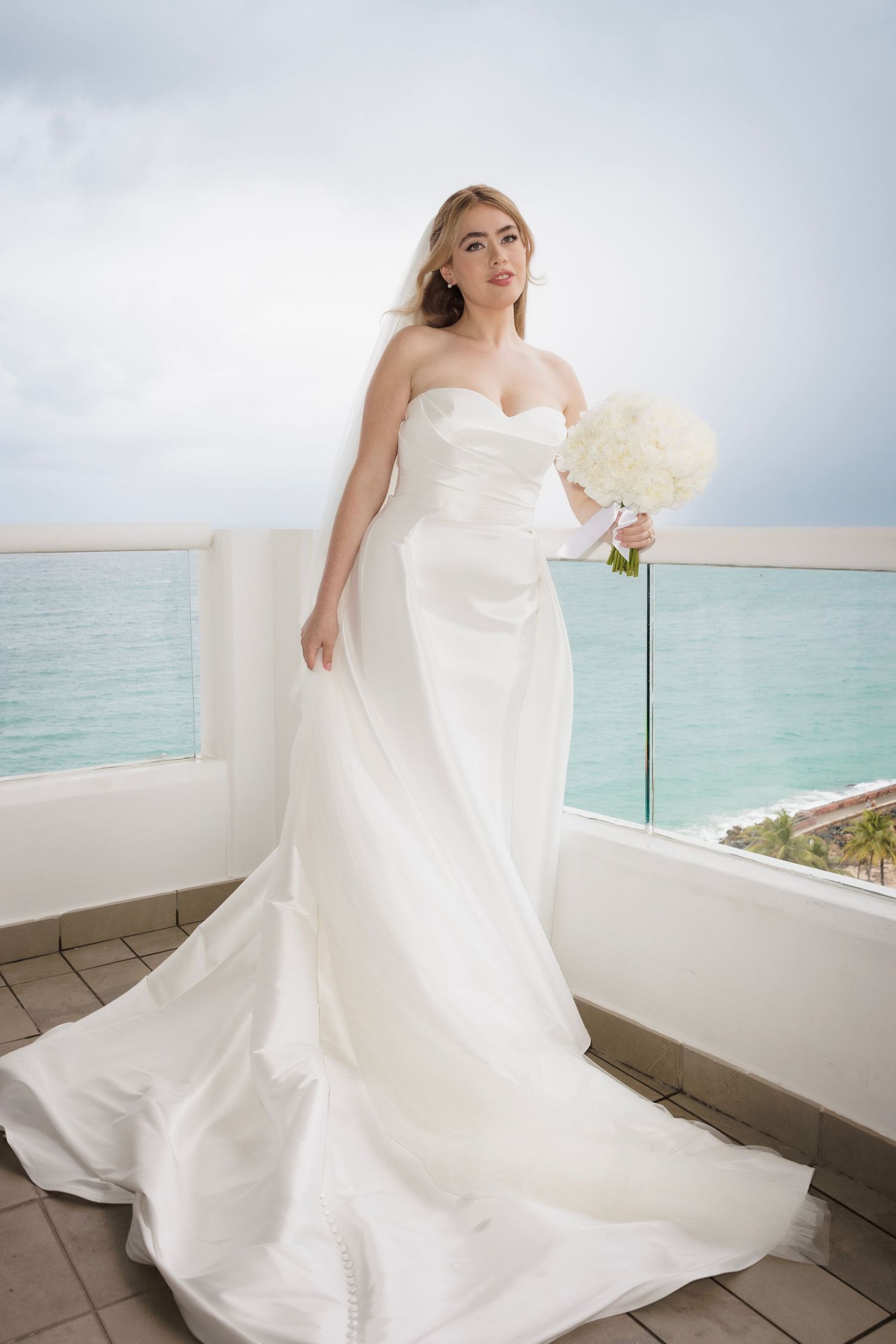 A bride in a flowing white wedding gown poses on an oceanfront balcony with turquoise waters in the background.