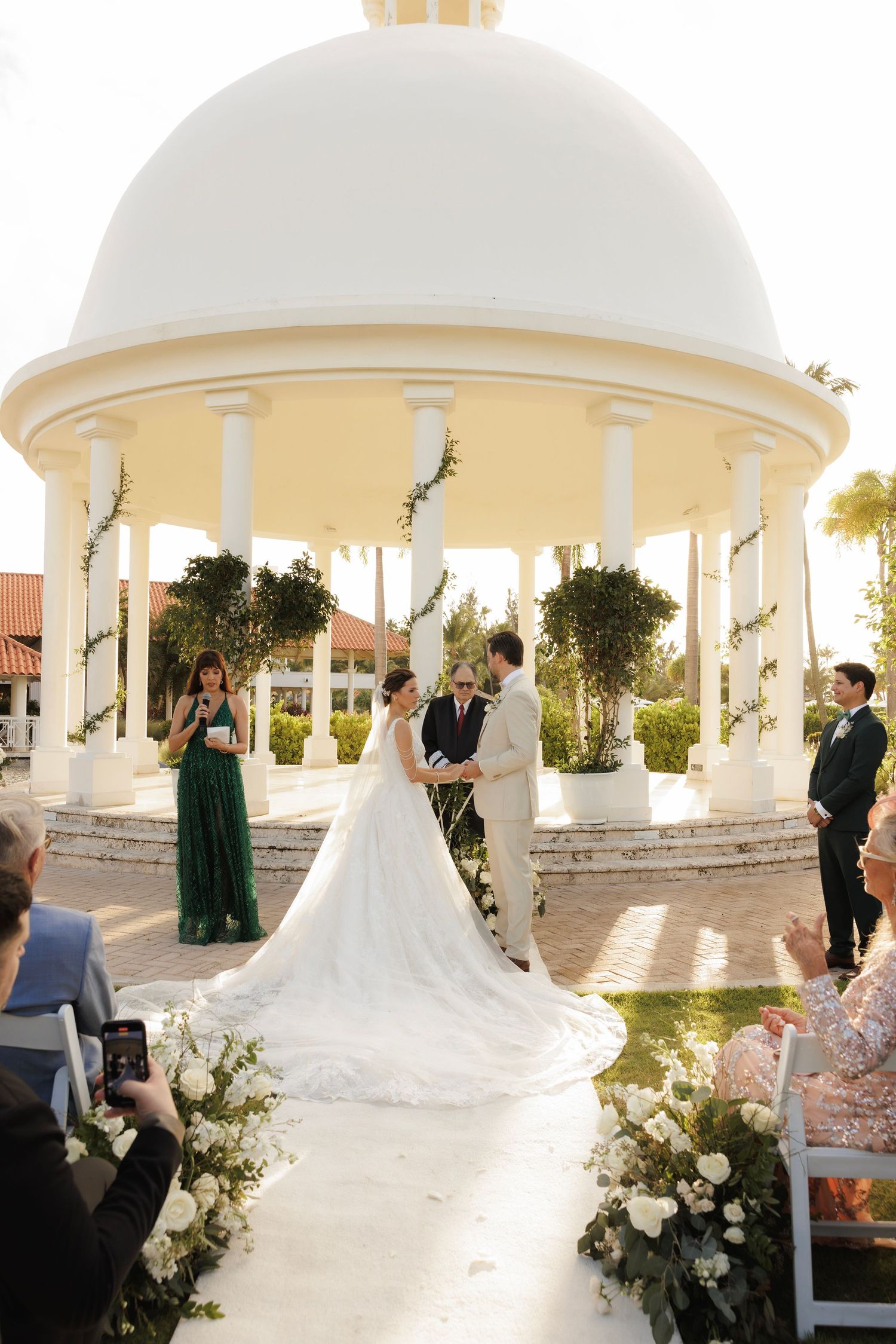 An elegant outdoor wedding ceremony takes place under a white gazebo with floral arrangements at sunset.