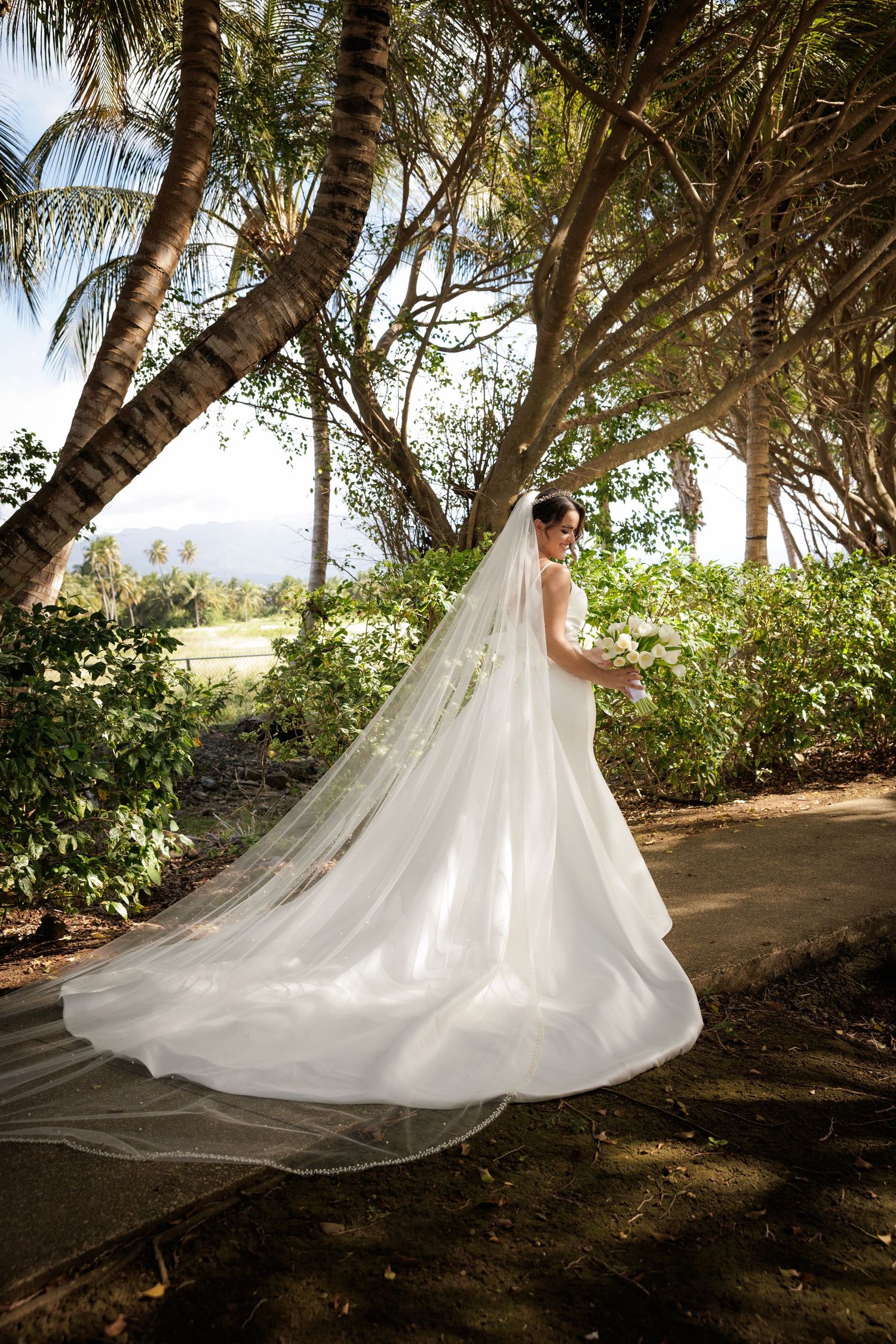 A stunning bride in a flowing white gown with a cathedral-length train poses among tropical palm trees on a beach.