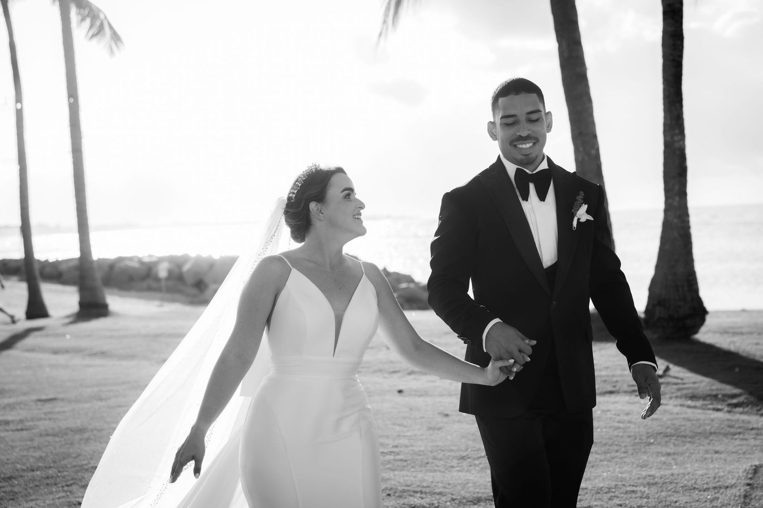Black and white photo of newlyweds joyfully walking together on a palm-lined path.