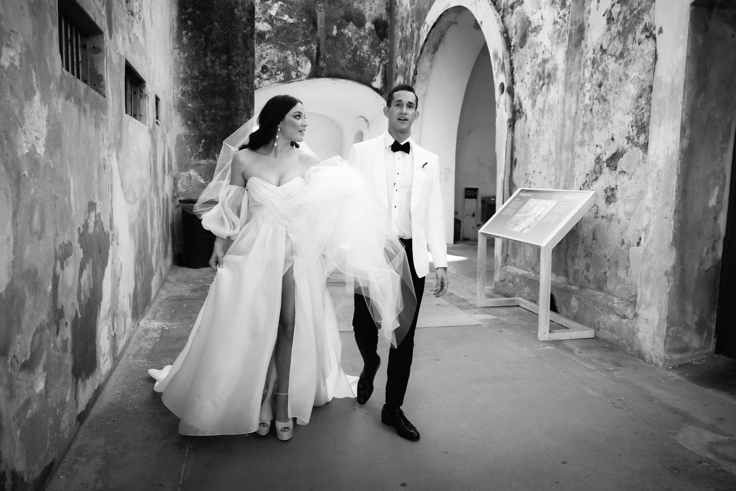 Newlyweds walk through historic stone passageway in formal wedding attire.