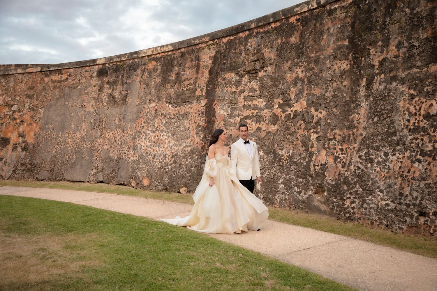 Wedding couple walking along ancient stone fortress walls under cloudy skies.