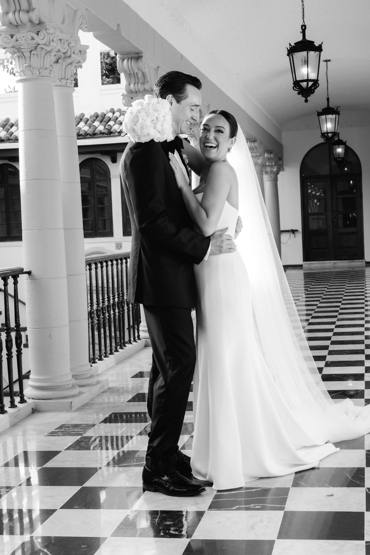 A couple shares a romantic moment in a stunning white-columned hallway with checkerboard flooring and elegant lanterns.
