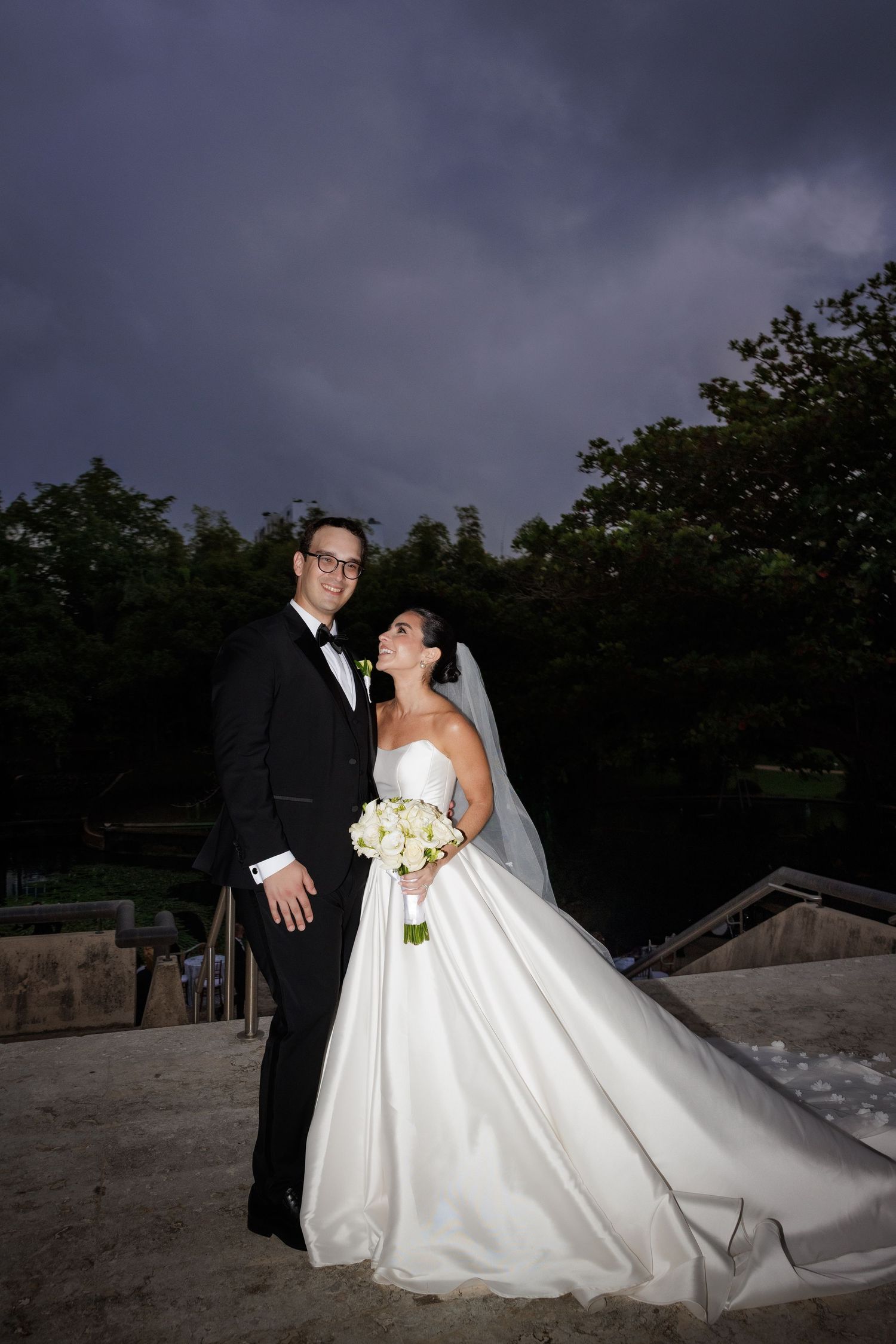A romantic wedding couple poses outdoors at dusk with dramatic stormy clouds in the background.