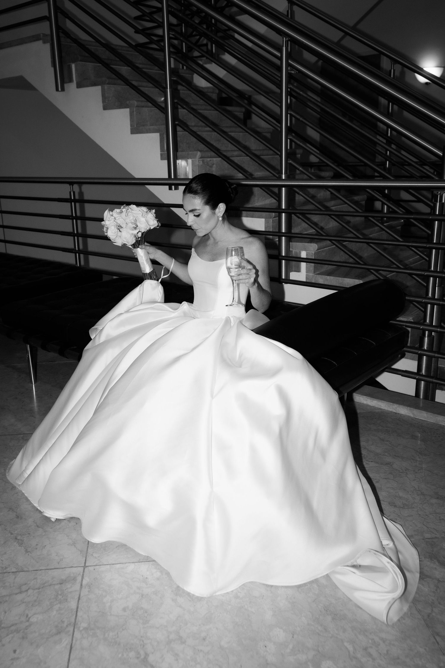 A bride in a flowing white ballgown poses dramatically on a modern staircase with a bouquet.