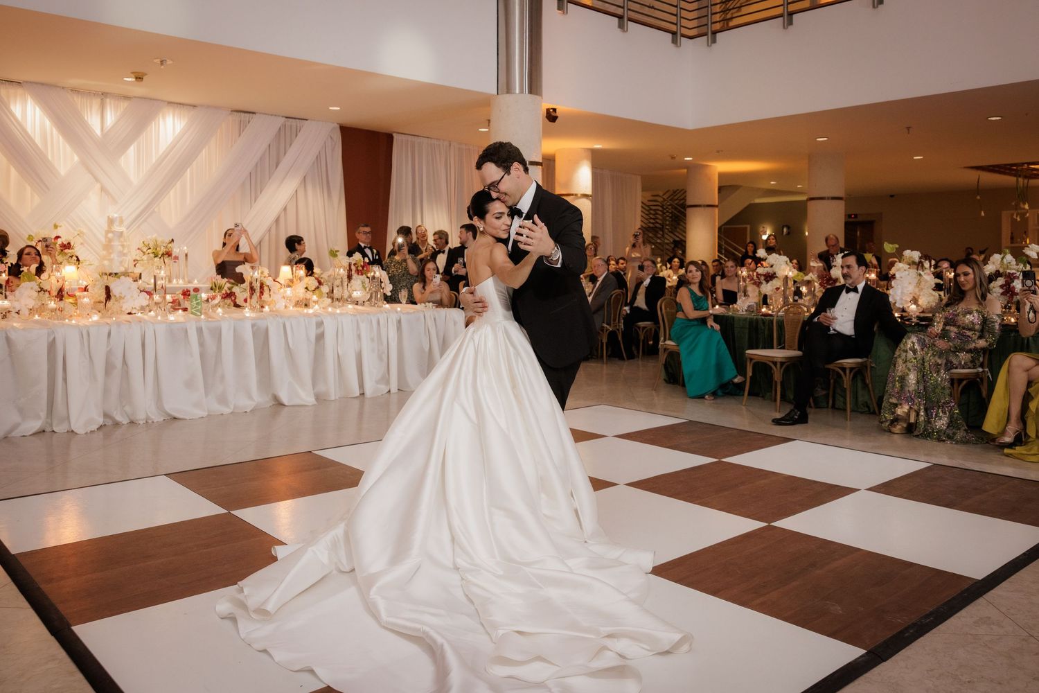 A wedding couple performs their first dance in a spacious ballroom surrounded by guests at their reception.