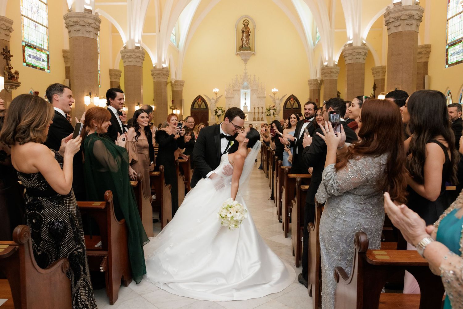 Wedding guests capture photos as newlyweds walk down the aisle in a beautifully decorated church.