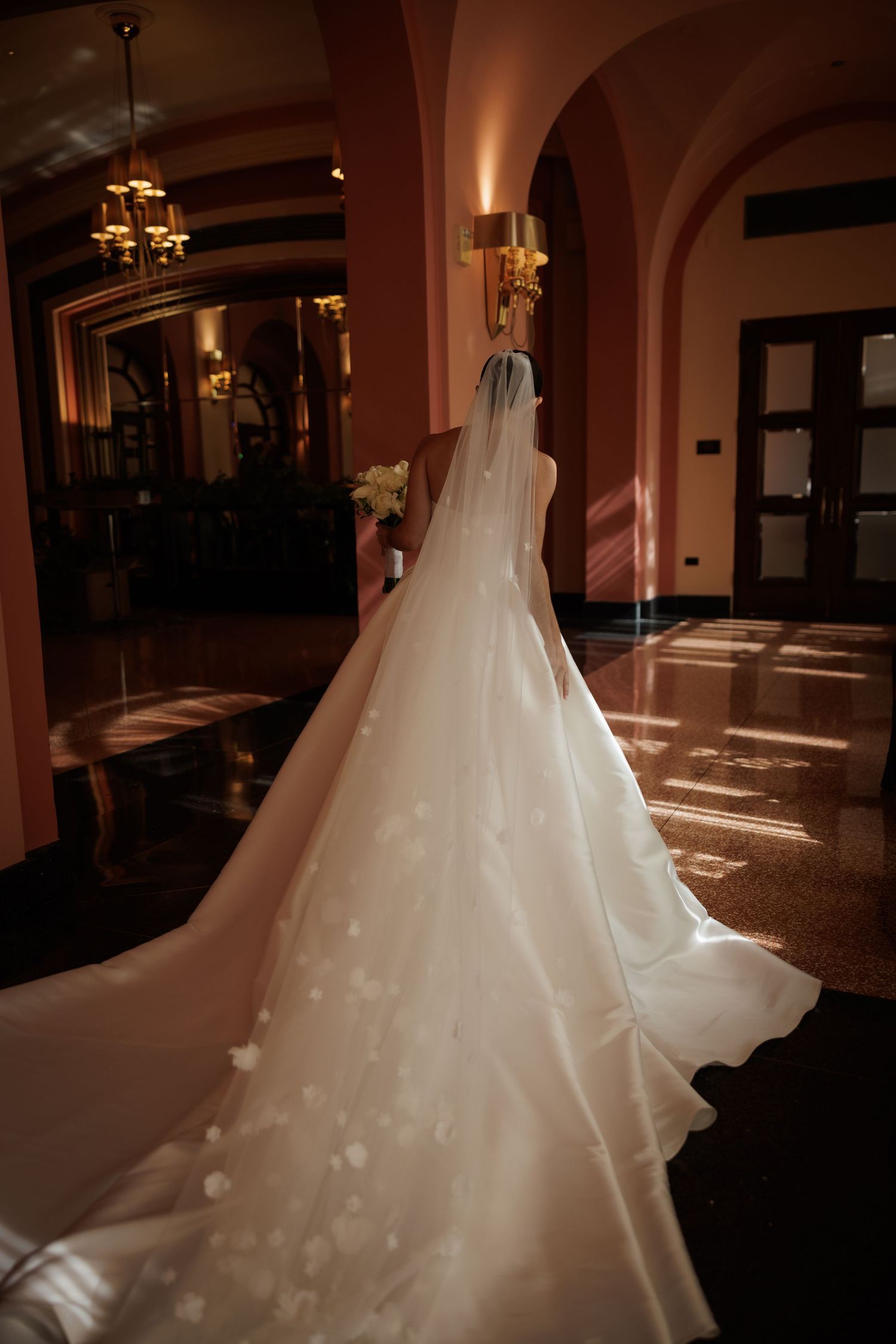 Flowing white wedding gown with long train photographed in dramatic lighting against dark hallway.