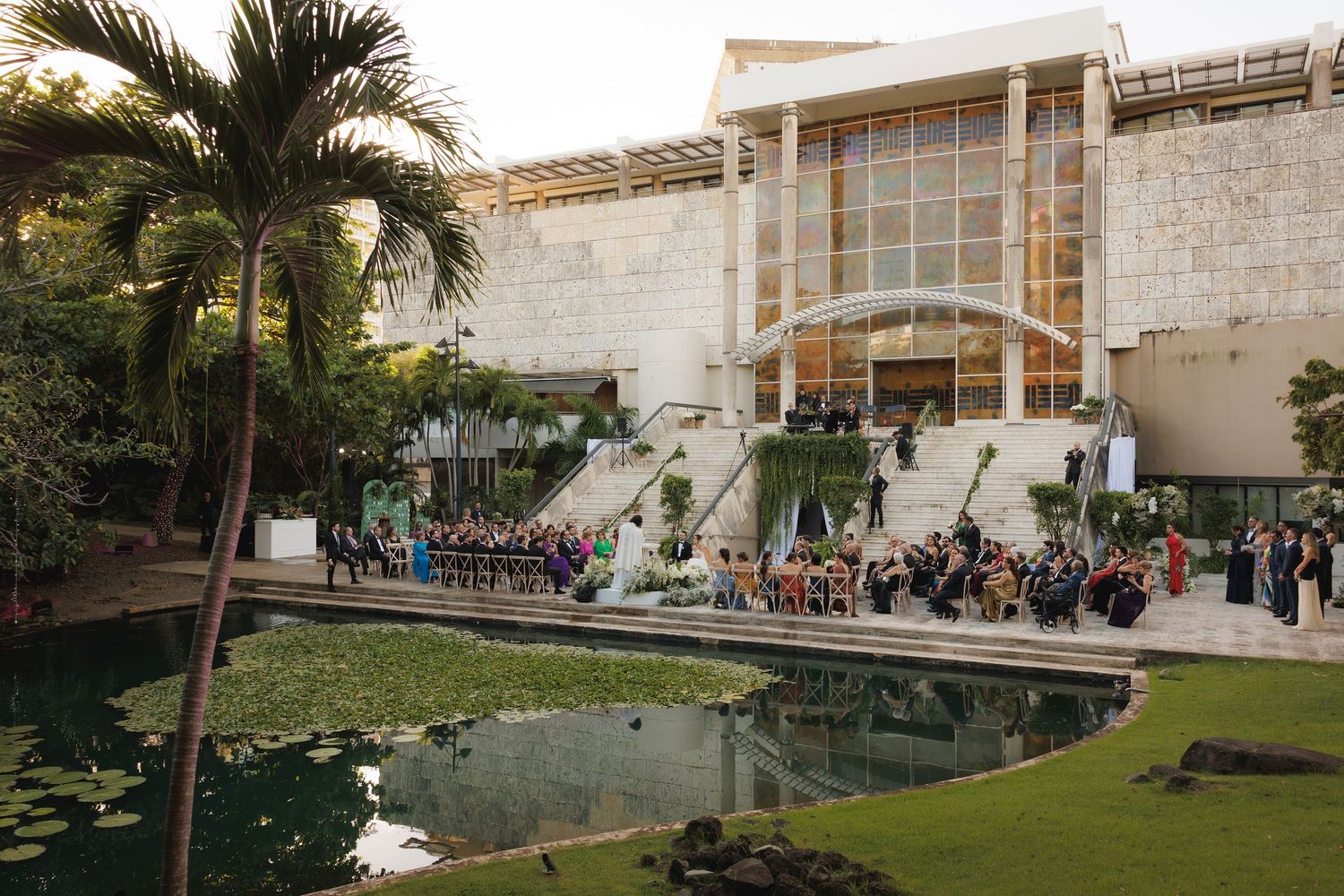 Modern stone venue with reflecting pool and palm trees hosting outdoor wedding ceremony with gathered guests.