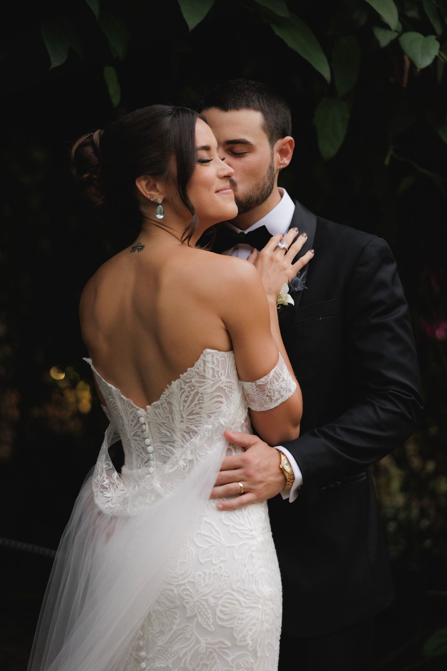 Bride in lace gown and groom share tender moments during their wedding photoshoot against dark foliage.