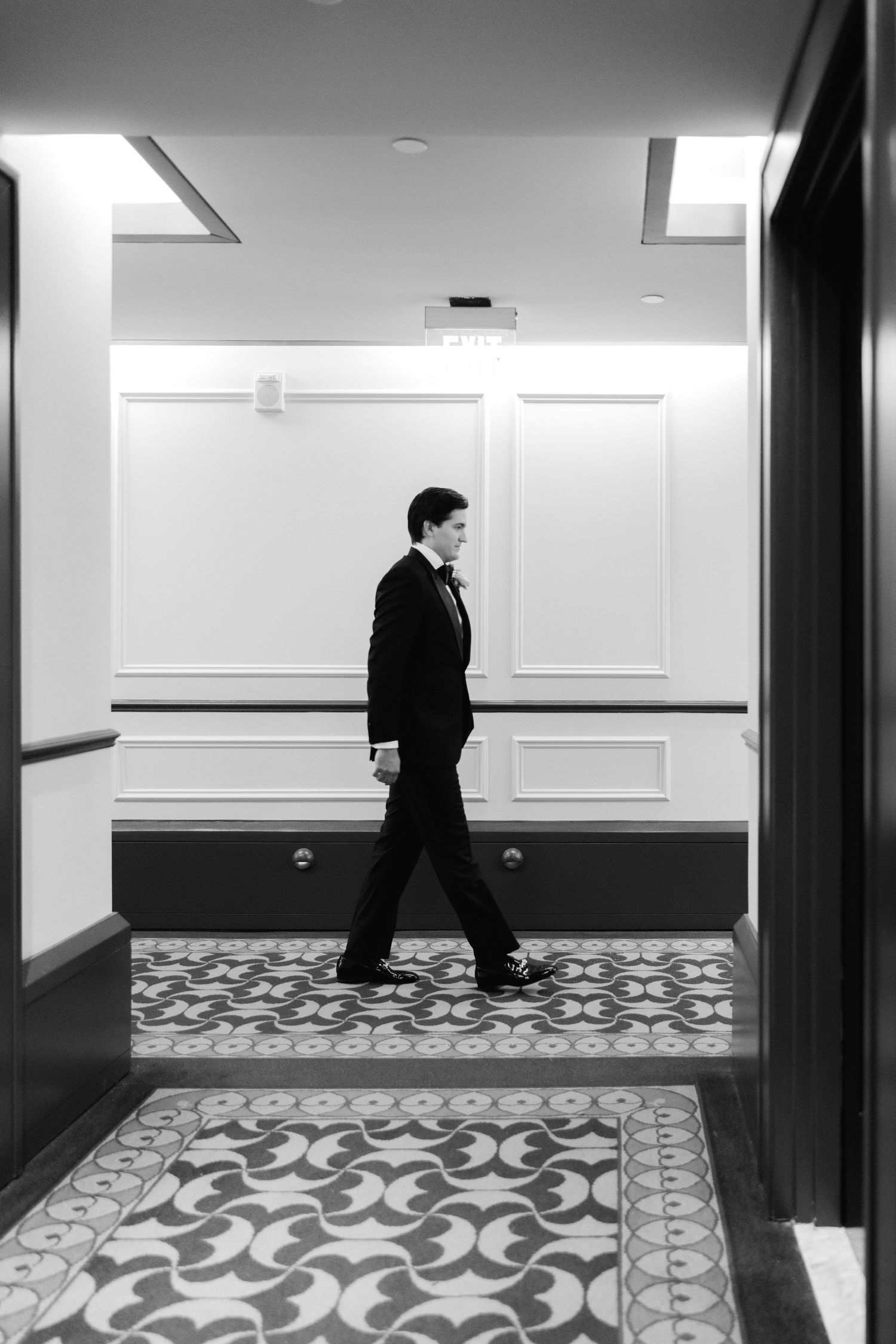 Minimalist black and white hallway photo showing figure walking past patterned carpet.