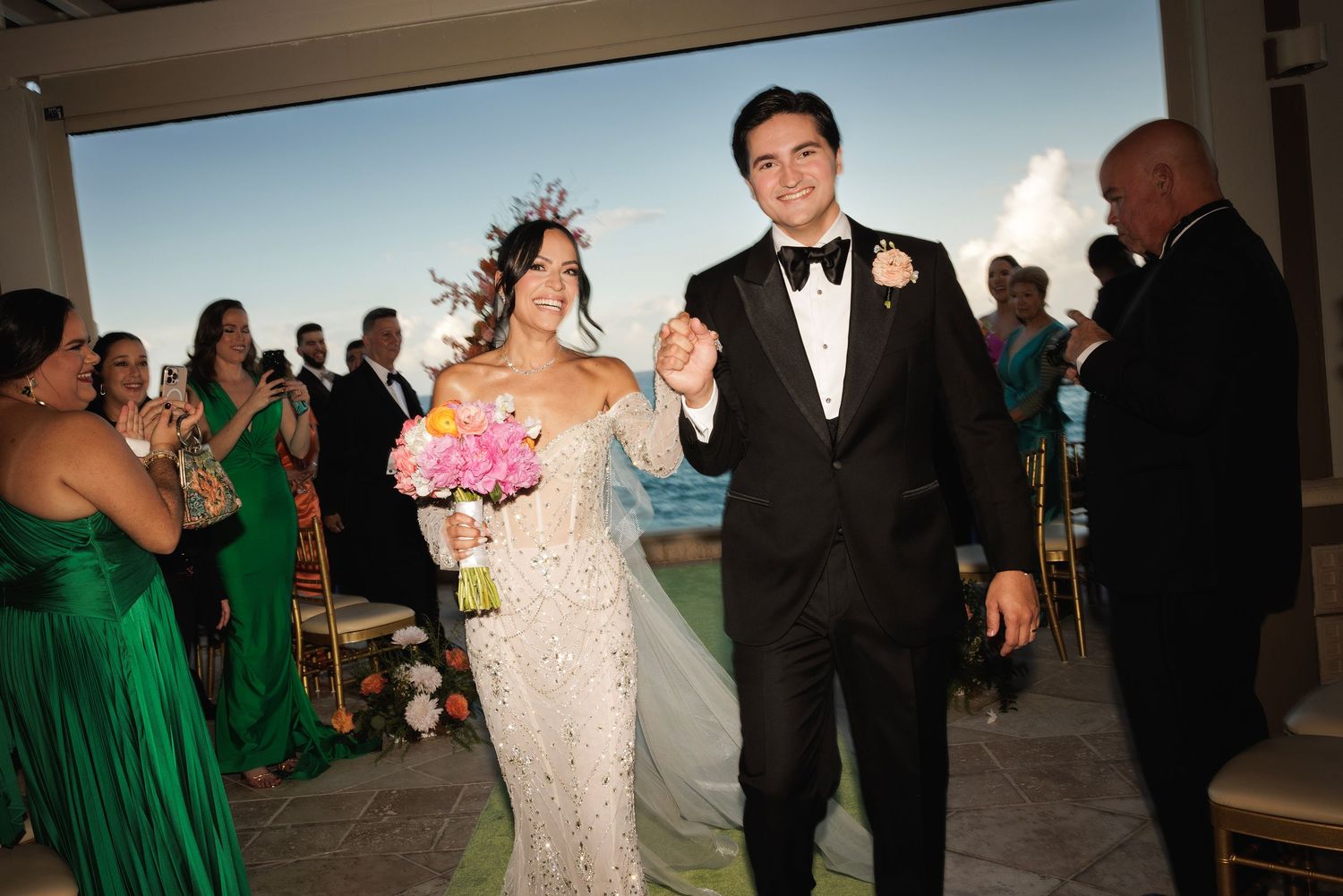 A joyful wedding couple walks down the aisle together while guests in emerald green dresses look on.