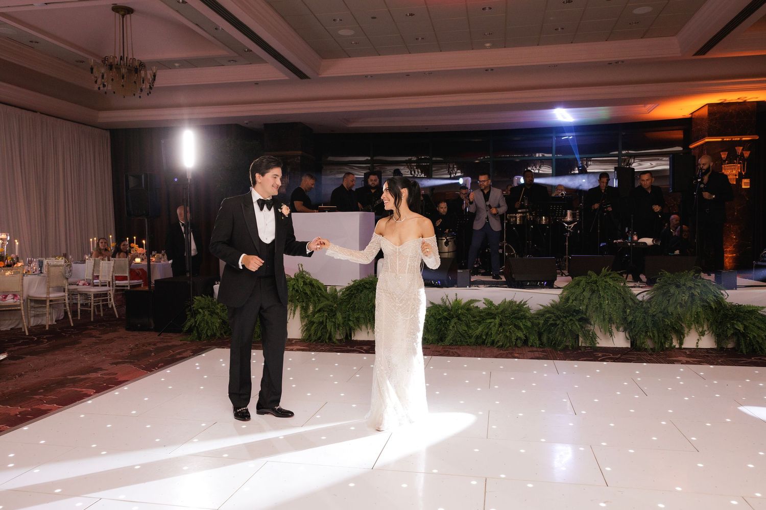 A couple stands together on an elegant white dance floor surrounded by ambient lighting at an indoor reception venue.