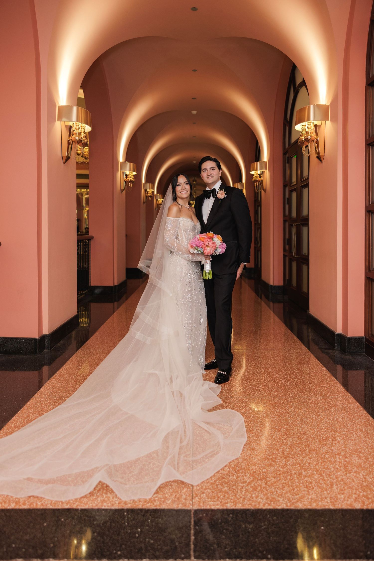 A wedding couple poses in an elegant pink arched hallway with ornate wall sconces and a flowing white bridal gown.