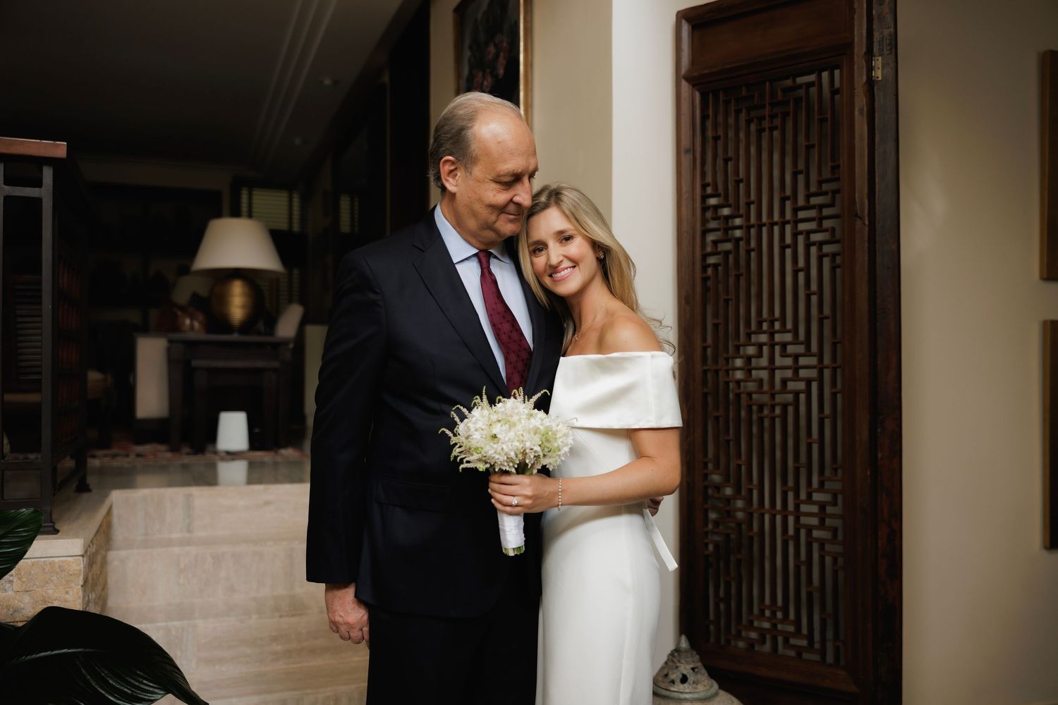A bride in an elegant white off-shoulder dress holds a white bouquet while posing inside an elegant home interior.